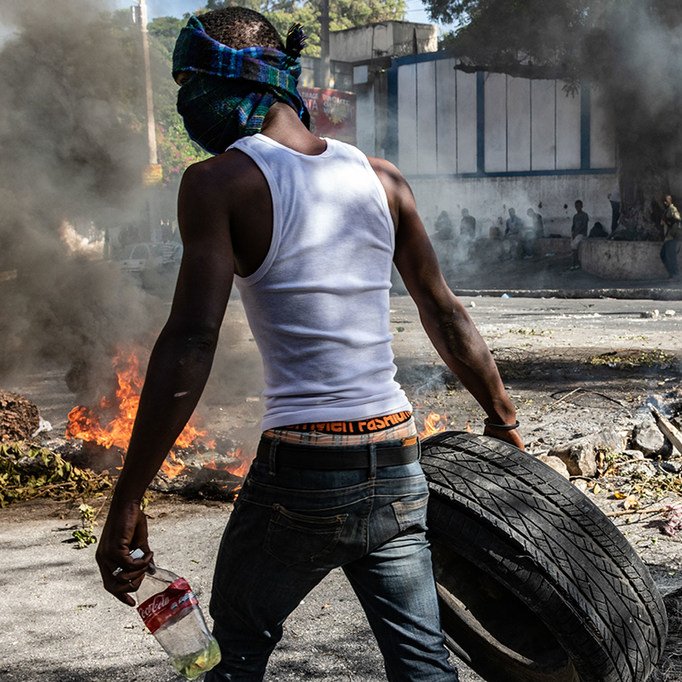 L'image montre une scène intense de protestation. Un homme, portant un débardeur blanc et un bandana sur le visage pour se protéger, est vu en arrière-plan. Il transporte un pneu que semble-t-il il va utiliser pour le jeter dans un feu. Autour de lui, on aperçoit de la fumée épaisse s'élevant dans l'air, résultant de plusieurs feux allumés sur la route. Le sol est jonché de débris, et l'atmosphère est chargée de tension, évoquant un sentiment de chaos et de lutte. Les flammes crépitent et laissent émaner une lumière orange. En arrière-plan, on peut distinguer des silhouettes de personnes, probablement d'autres manifestants.