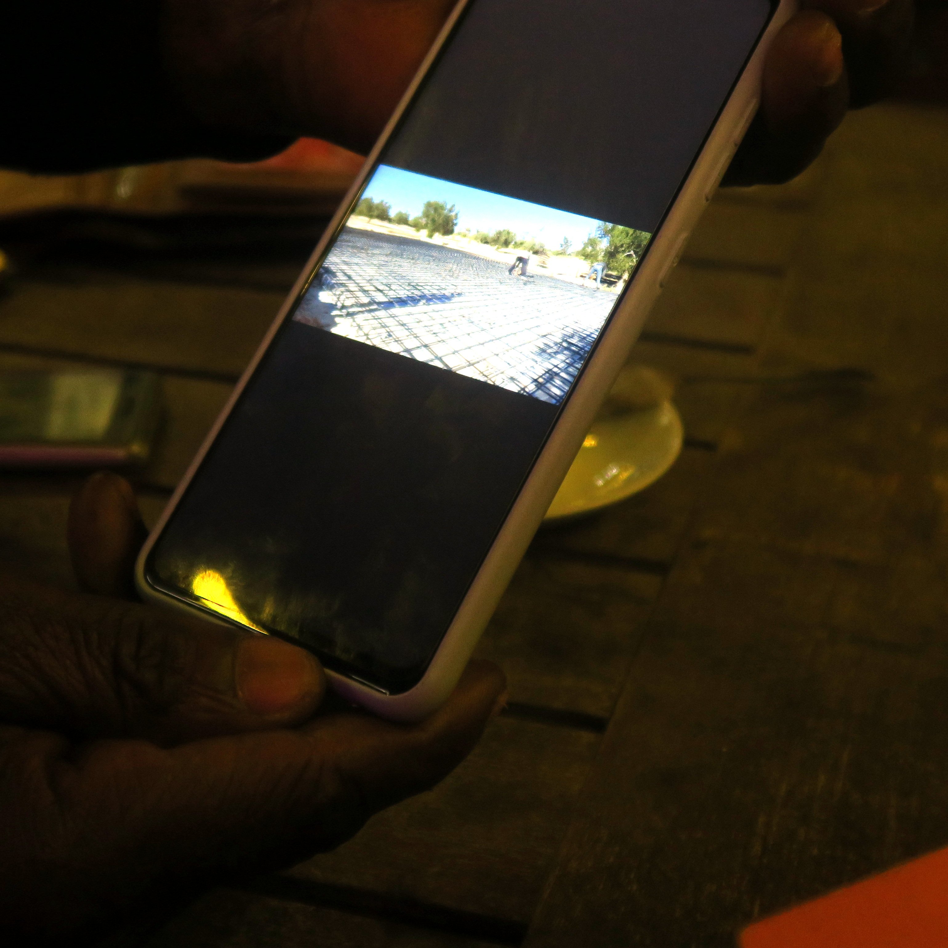 L'image montre une main tenant un téléphone portable, qui affiche une photographiée (ou une vidéo) d'un paysage. À l'arrière-plan, on peut imaginer une scène en extérieur, probablement un terrain ou une route. La lumière dans la pièce est tamisée, et il y a une table en bois visible. Sur la table, on peut apercevoir quelques objets, comme un petit paquet de cigarettes et un plateau. L'ambiance semble tranquille et décontractée, suggérant un moment de détente ou de partage.