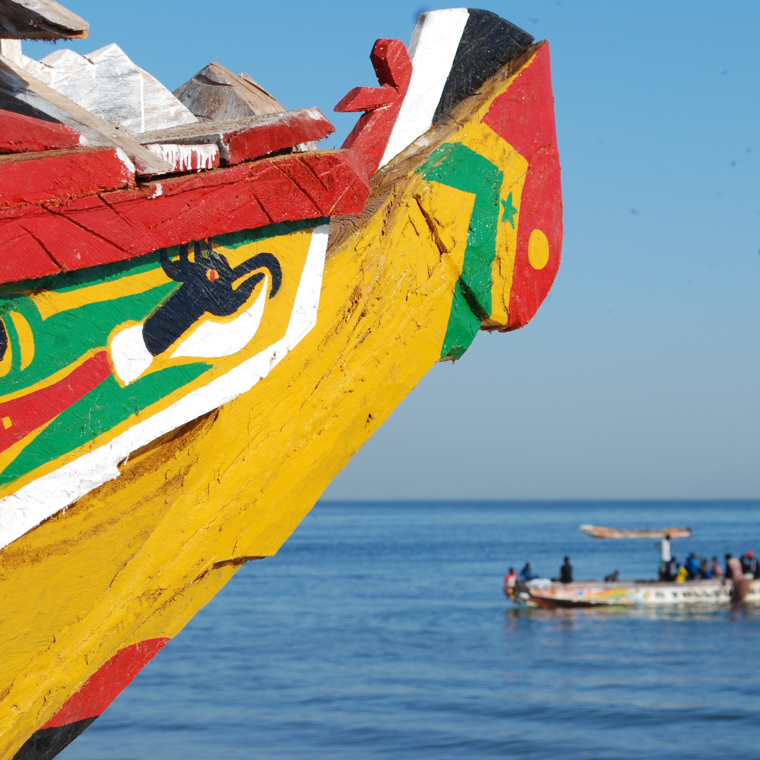 Cette image représente la proue colorée d'une pirogue traditionnelle, peinte de motifs vifs dans des couleurs comme le rouge, le jaune, et le vert. Au loin, on peut apercevoir d'autres bateaux de pêche qui naviguent paisiblement sur l'eau calme d'une mer bleue. Le ciel est dégagé, créant une atmosphère sereine et ensoleillée. L'air salin doit être frais, et on peut entendre le bruit des vagues et peut-être des conversations des marins qui travaillent sur leurs embarcations. L'ensemble dégage une ambiance maritime typique des côtes africaines.
