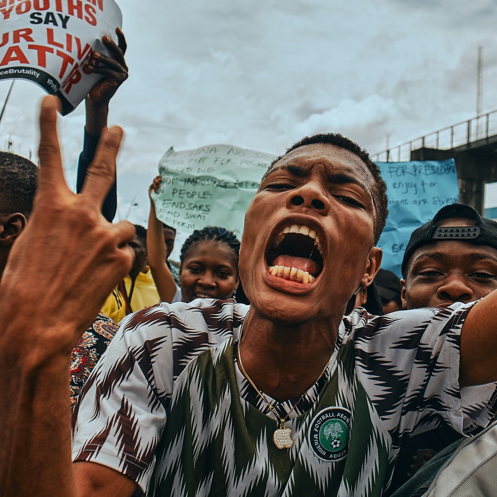 L'image montre une scène de manifestation animée. Au premier plan, un jeune homme crie avec passion, levant les deux doigts en signe de victoire ou de protestation. Son visage exprime une forte intensité émotionnelle, avec une bouche ouverte et des yeux déterminés. Il porte un maillot à motifs verts et blancs, représentant probablement les couleurs de son pays. Derrière lui, un groupe de personnes brandit des pancartes avec des slogans, illustrant un sentiment de mobilisation et de solidarité. L'ambiance est vibrante, évoquant une lutte collective pour des causes qui leur tiennent à cœur, sous un ciel nuageux.