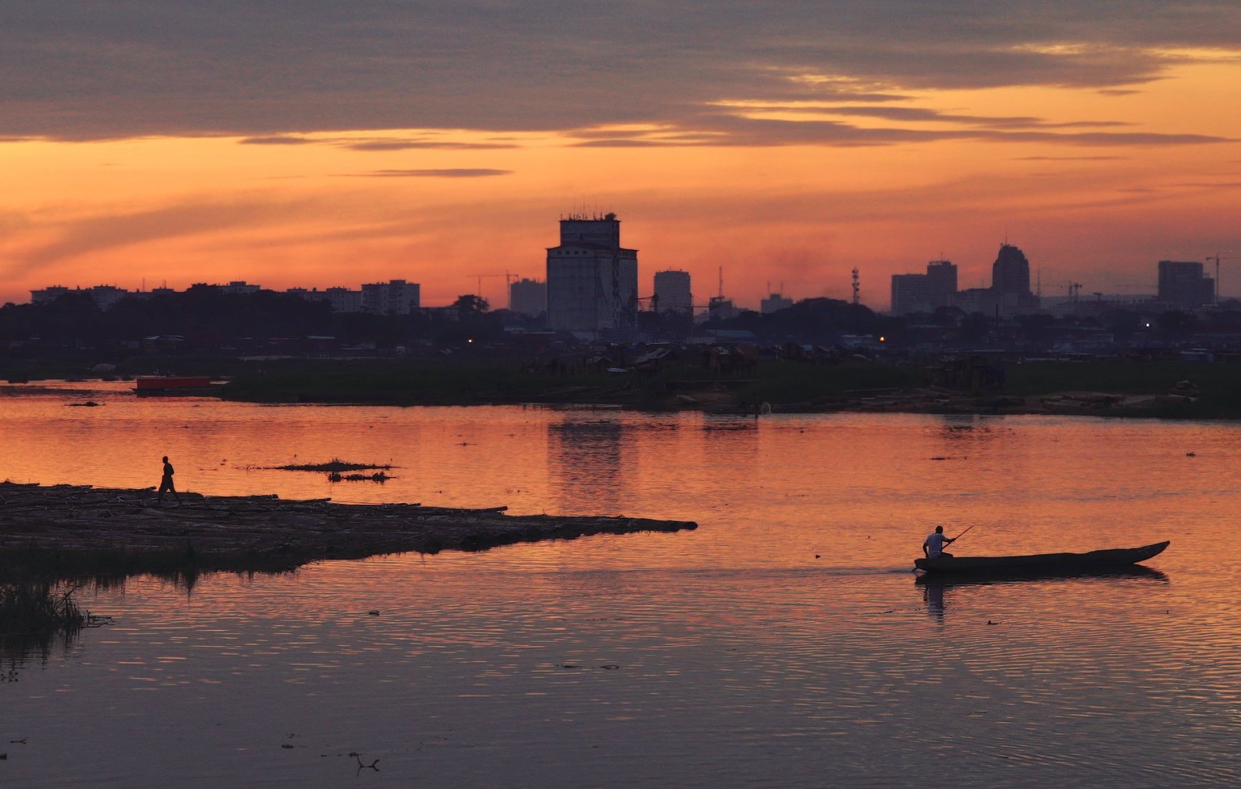 L'image dépeint un paysage paisible au crépuscule. Le ciel est teinté de nuances chaudes d'orange, de rose et de violet, créant une atmosphère calme et sereine. À l'avant, un homme marche au bord de l'eau, tandis qu'un autre navigue tranquillement sur un petit bateau. La surface de la rivière reflète les couleurs du ciel, ajoutant à la beauté de la scène. En arrière-plan, on aperçoit des silhouettes de bâtiments urbains, suggérant la proximité d'une ville. L'ensemble évoque une sensation de tranquillité et de connexion avec la nature, tout en étant en harmonie avec l'activité humaine.
