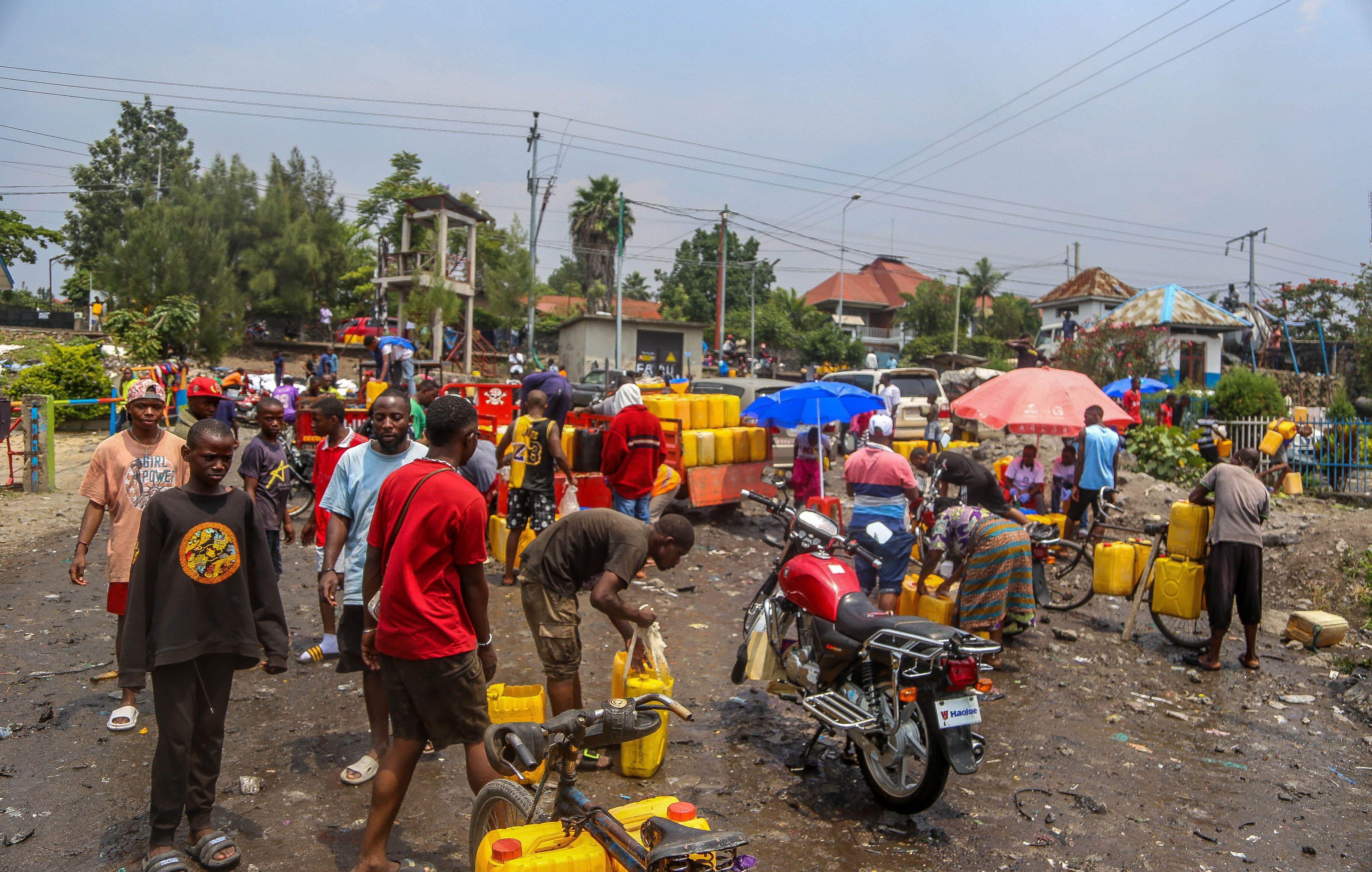 The image depicts a crowded scene in an outdoor setting, likely a market or gathering place. People are seen collecting water from yellow jerry cans, some of which are stacked nearby. The atmosphere appears busy and somewhat chaotic, with individuals interacting and engaging in various activities. There are motorcycles and umbrellas visible, suggesting a warm, sunny environment. The ground is littered with debris, indicating a lack of cleanliness in the area. The background features buildings and trees, contributing to the urban landscape.