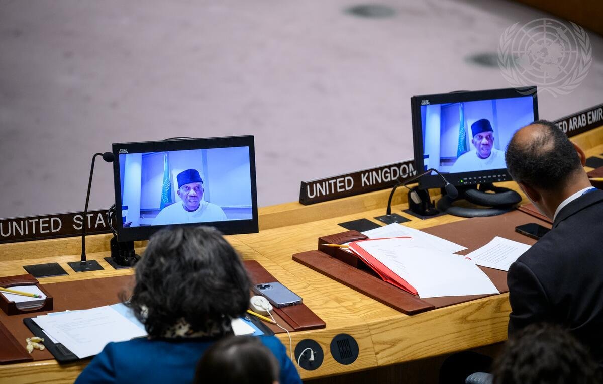 L'image montre une scène à l'intérieur d'une salle de conférence, probablement lors d'une réunion d'une organisation internationale comme l'ONU. Au premier plan, deux personnes sont assises à une grande table en bois, devant des ordinateurs qui affichent des visages sur écran. L'un des intervenants porte un couvre-chef traditionnel. On devine qu'ils sont en train d'assister à une vidéoconférence. Sur la table, il y a des documents empilés, des fournitures de bureau et des éléments d'identification des pays présents, comme ceux des États-Unis et du Royaume-Uni. L'atmosphère est formelle, accentuée par la présence de symboles et d'éléments décoratifs liés à l'organisation.