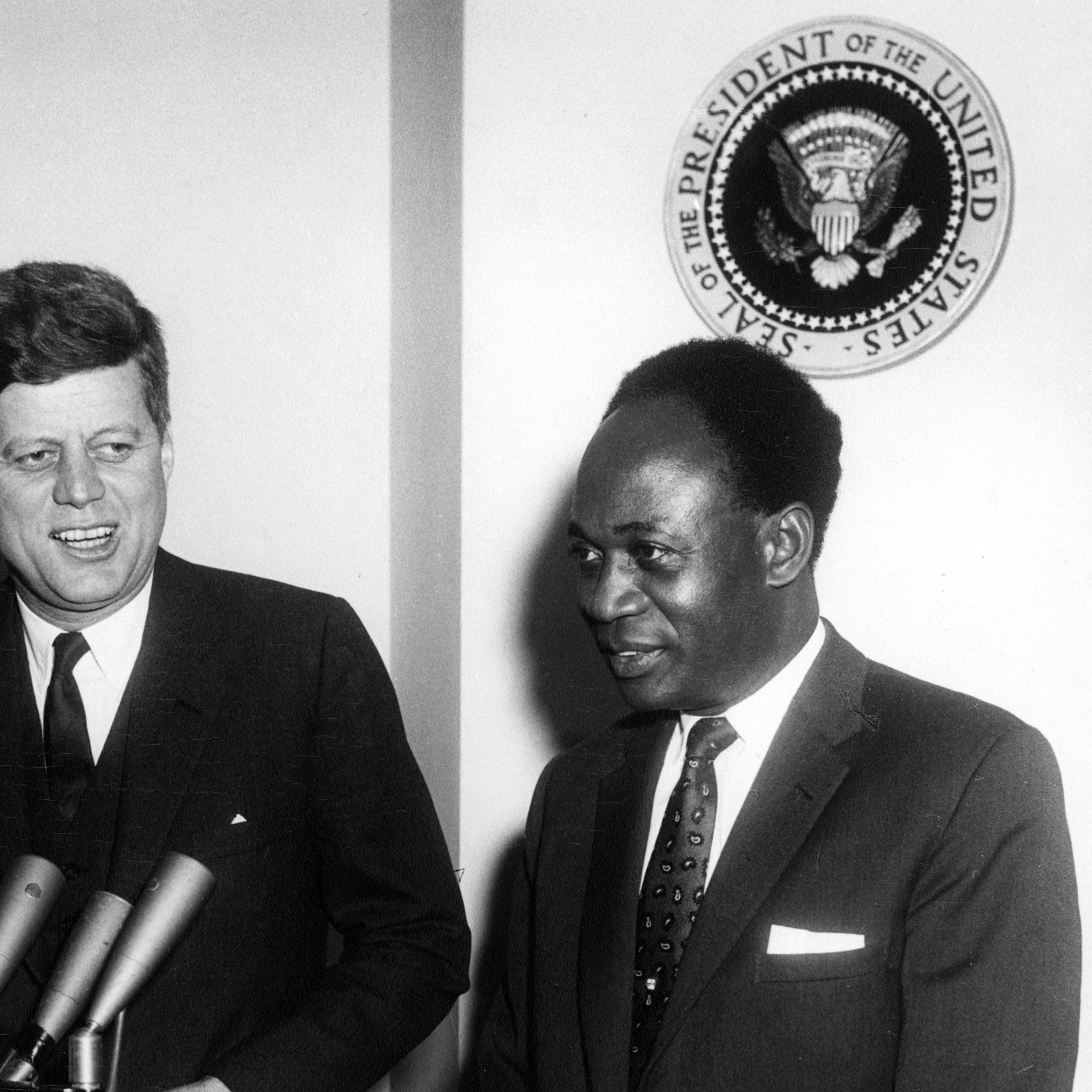 The image features two men, one of whom is John F. Kennedy, the President of the United States, and the other is likely Kwame Nkrumah, the first President of Ghana. They appear to be at a press conference or a formal meeting, with microphones in front of them. Behind them, there is a seal that represents the office of the President of the United States. Both men are dressed formally, with Kennedy in a suit and tie, and they are smiling, suggesting a cordial atmosphere.
