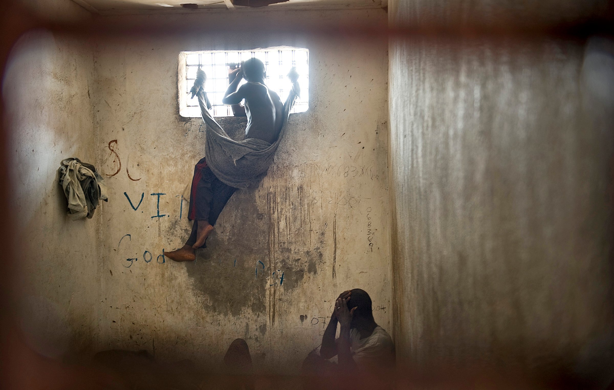 L'image dépeint un environnement sombre et restreint, vraisemblablement une cellule ou une pièce fermée. Au premier plan, deux personnes sont présentes. L'une est assise, visiblement accroupie, la tête entre les mains, adoptant une posture de détresse ou de réflexion. L'autre personne est perchée près d'une fenêtre grillagée, les jambes repliées, enveloppée dans un drap qui semble être une couverture. La lumière qui pénètre par la fenêtre est limitée, créant un contraste entre l'extérieur et l'intérieur sombre de la pièce. Les murs portent des marques, probablement des graffiti, ce qui suggère un environnement négligé. L'atmosphère générale est lourde et empreinte de mélancolie, reflétant un sentiment d'isolement et de désespoir.