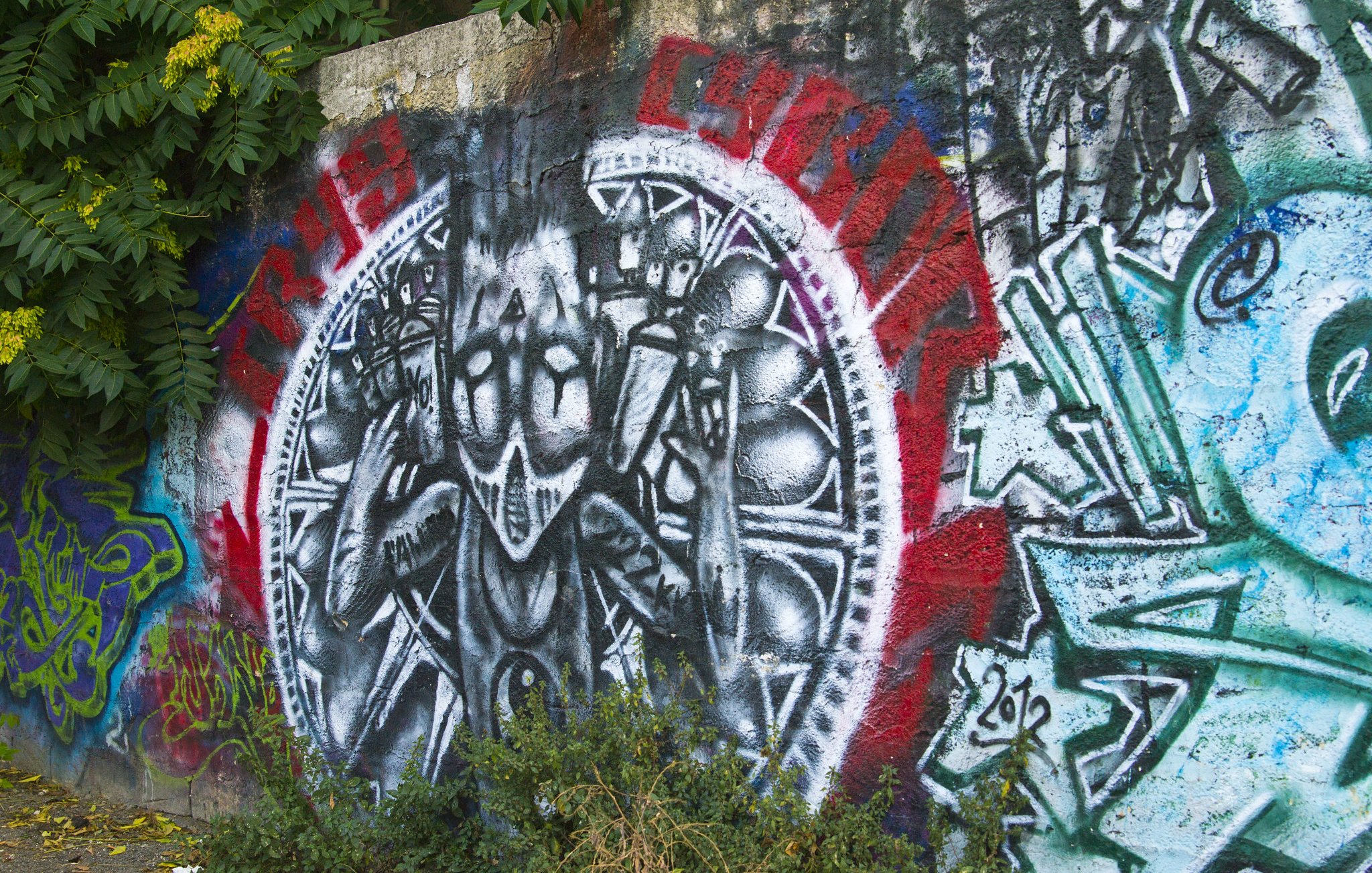 L'image représente une peinture murale vibrante sur un mur en béton. Au centre, on peut voir une figure stylisée, habillée de manière étrange, qui semble exprimer une sorte de défi ou de résistance avec des gestes des mains. La figure est entourée d'un motif circulaire qui est accentué par des couleurs vives comme le rouge et le noir, créant un fort contraste visuel. D'autres graffitis colorés sont visibles autour, ajoutant une diversité de formes et de couleurs à l'ensemble. À la base, des feuilles vertes, provenant de la végétation environnante, sortent du sol, créant une atmosphère vivante et urbaine.