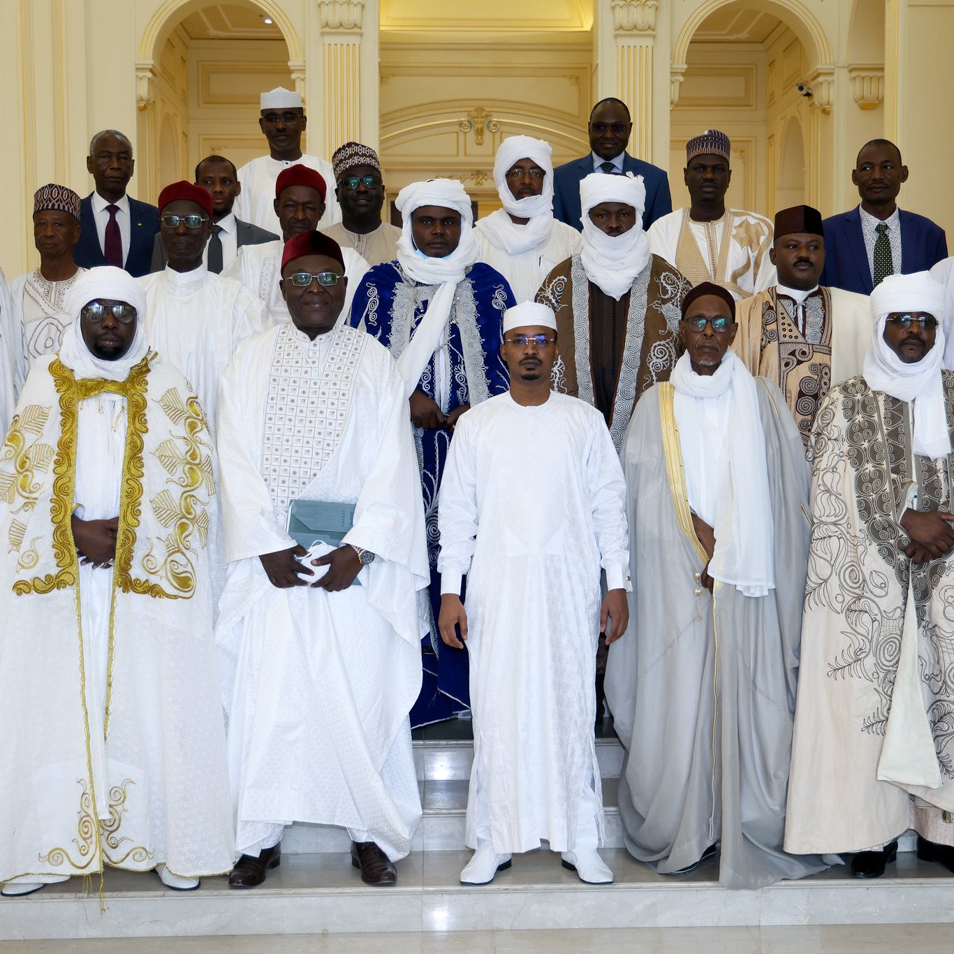 L'image montre un groupe de personnes habillées en vêtements traditionnels, rassemblées dans un espace élégant. Ils sont principalement vêtus de robes blanches, avec des éléments colorés comme des capes vertes et bleues. Les hommes portent des turbans et des chapeaux, ajoutant à la richesse de leurs tenues. L'arrière-plan est un intérieur lumineux et spacieux, avec des colonnes et des décorations architecturales. Le groupe semble coordonné et solennel, probablement pour une occasion spéciale.
