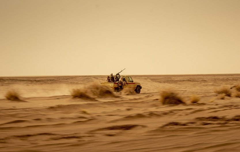 L'image montre un véhicule tout-terrain, probablement un pick-up, qui avance rapidement à travers un désert. La poussière s'élève autour de lui, créant une ambiance de mouvement et de vitesse. À l'arrière du véhicule, on aperçoit des silhouettes de personnes, probablement des passagers, qui semblent être équipés pour un environnement hostile. Le paysage est vaste et aride, avec des dunes de sable et des buissons résistant aux conditions sèches. Le ciel est clair mais teinté d'une couleur jaunâtre, suggérant une chaleur intense. L'ensemble donne une impression de solitude et d'aventure dans un milieu difficile.