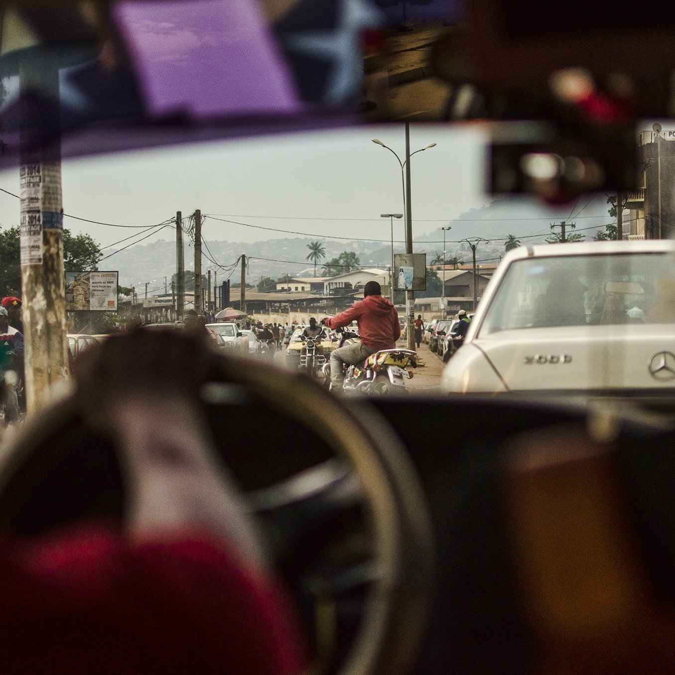 L'image présente une scène urbaine animée vue depuis l'intérieur d'un véhicule. On peut voir le côté d'un tableau de bord, avec une main tenant le volant. À travers le pare-brise, la circulation est dense, comprenant des voitures et des motos. On distingue une personne à vélo sur la route, et au fond, des bâtiments et des montagnes se dessinent à l'horizon. L'atmosphère semble vivante et mouvementée, caractéristique de la vie citadine. La lumière du jour éclaire la scène, révélant des détails sur les véhicules et les personnes qui circulent.