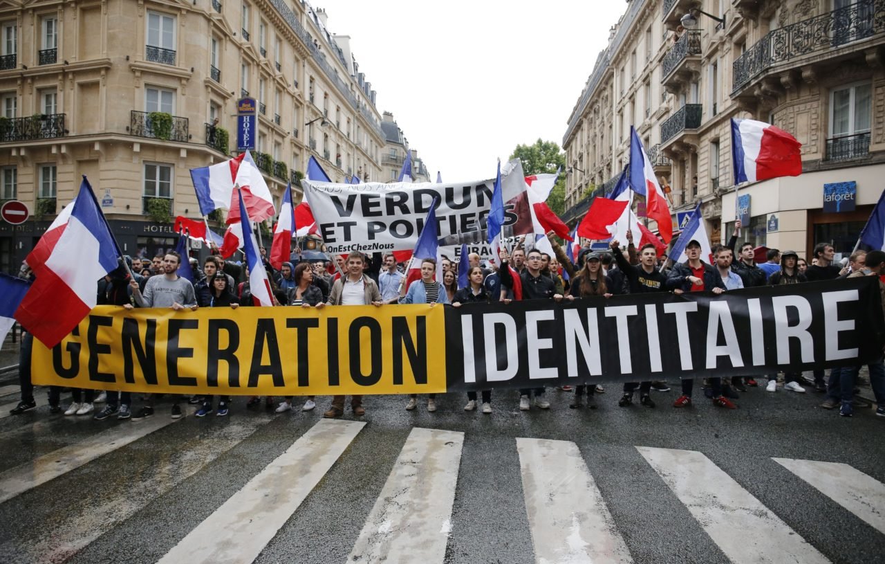 L'image montre une manifestation dans une rue urbaine. Un groupe de personnes tient une grande banderole noire avec le texte "GENERATION IDENTITAIRE" en lettres jaunes bien visibles. Elles brandissent également de nombreux drapeaux tricolores français, qui flottent au-dessus d'elles. En arrière-plan, on aperçoit une autre bannière avec les mots "VERDUN ET POITERS", indiquant probablement des messages ou des revendications relatives à ces lieux. Les manifestants semblent unis et déterminés, marchant ensemble sur le passage piéton, tandis que des bâtiments typiques de la ville se dressent à leurs côtés. L'ambiance générale est celle d'une démonstration publique, avec une forte présence de symboles nationaux.