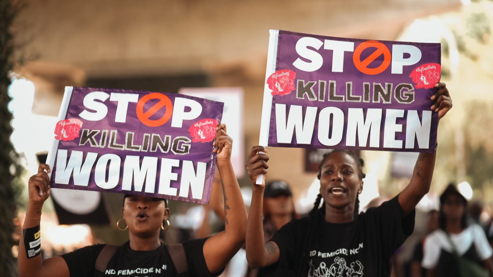 L'image montre deux femmes participante à une manifestation. Elles tiennent chacune une grande pancarte. Les pancartes portent le message "STOP KILLING WOMEN" en lettres capitales de couleur blanche, avec un symbole barré en rouge. Les femmes ont des expressions déterminées et sont vêtues de t-shirts noirs. L'arrière-plan suggère une ambiance de mobilisation avec d'autres personnes qui marchent et participent à l'événement. L'atmosphère est chargée d'émotion et de solidarité.