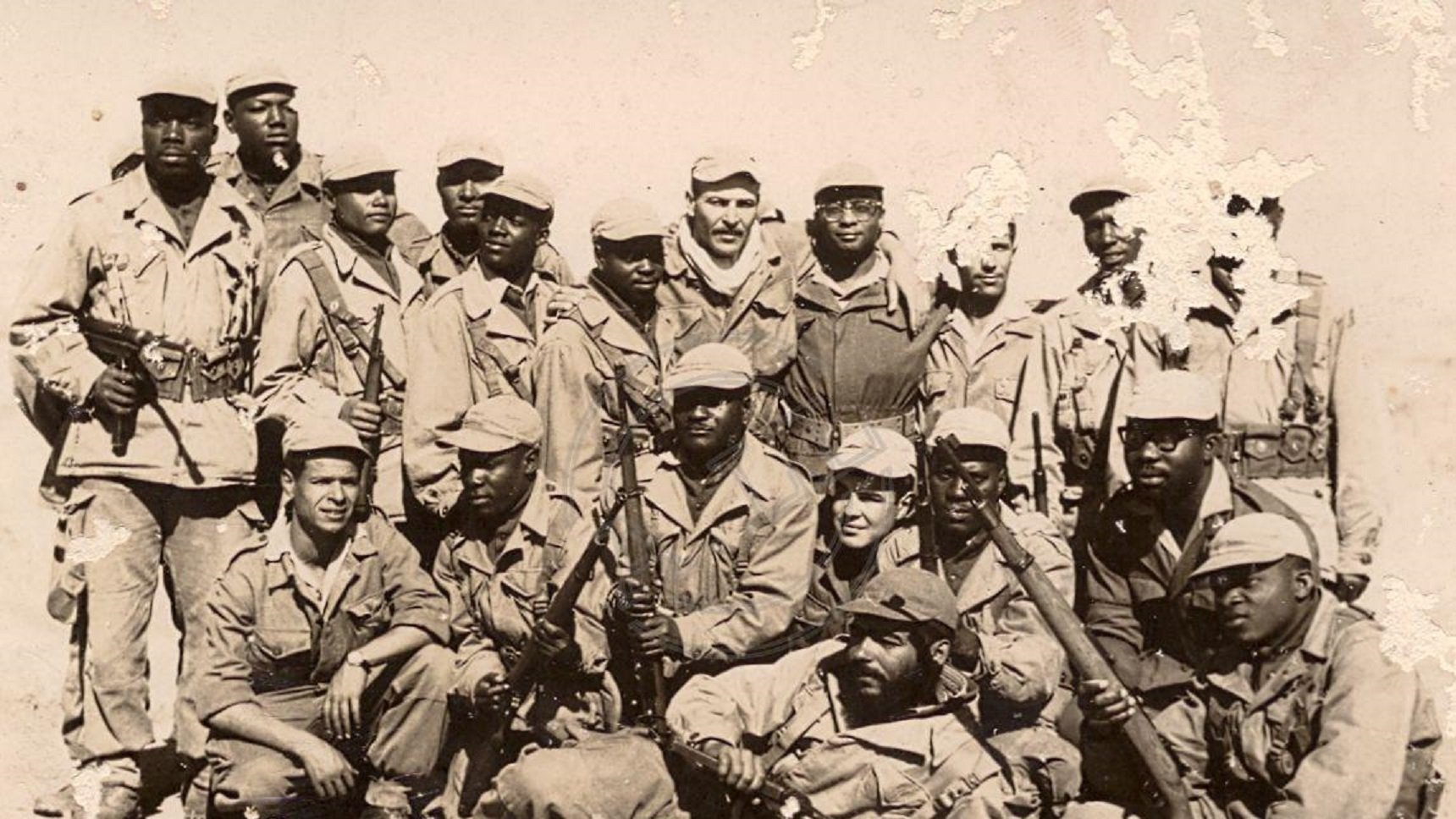 Cette image montre un groupe de soldats, probablement dans un contexte historique. Ils sont vêtus d'uniformes militaires, la plupart portant des chapeaux ou casquettes. Certains d'entre eux tiennent des armes, et l'ambiance générale dégage un sentiment de camaraderie et de force. Les soldats semblent être regroupés, avec une variation dans leurs postures : certains se tiennent debout tandis que d'autres sont accroupis ou assis. Le fond de la photo est déteint, ce qui suggère qu'il pourrait s'agir d'une image ancienne. L'ensemble de cette scène évoque un moment de solidarité et de détermination au sein du groupe.