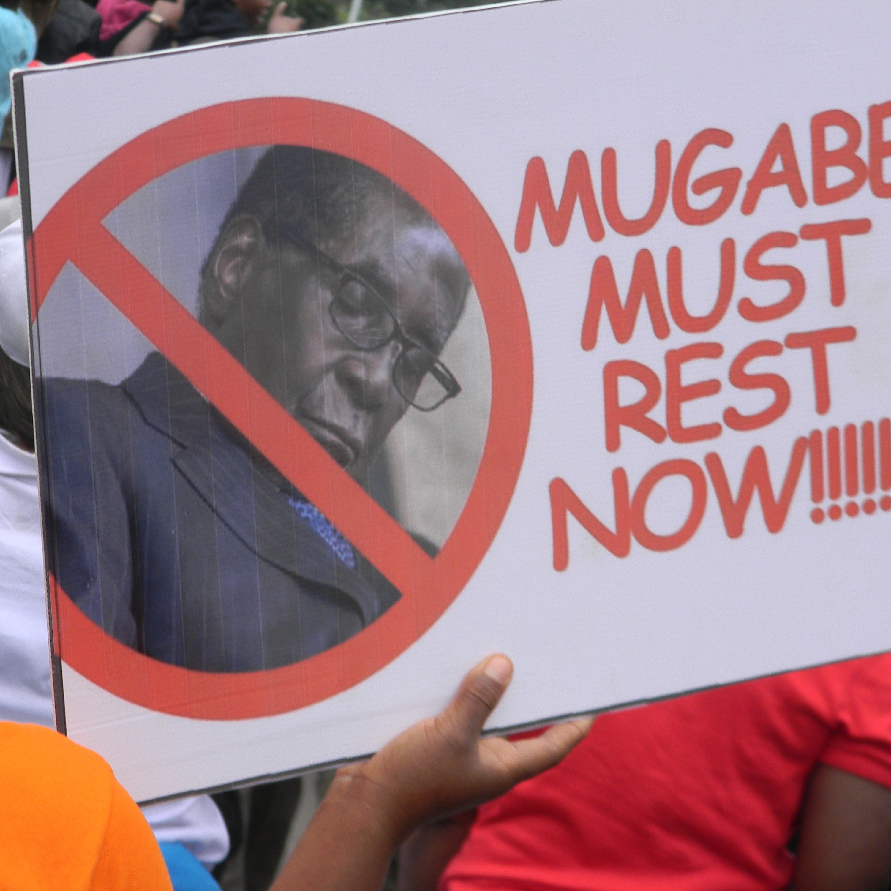 Cette image montre une personne tenant une pancarte lors d'une manifestation. La pancarte arbore un grand symbole barré, avec une photo d'un homme, généralement associé à un discours politique. Le texte sur la pancarte se lit : "MUGABE MUST REST NOW", exprimant un message clair de résistance ou de demande de cessation à l'égard de cette personne. En arrière-plan, on peut voir une foule, suggérant un rassemblement où des gens partagent des opinions fortes sur un sujet d'actualité. L'ambiance générale semble être celle d'une protestation.