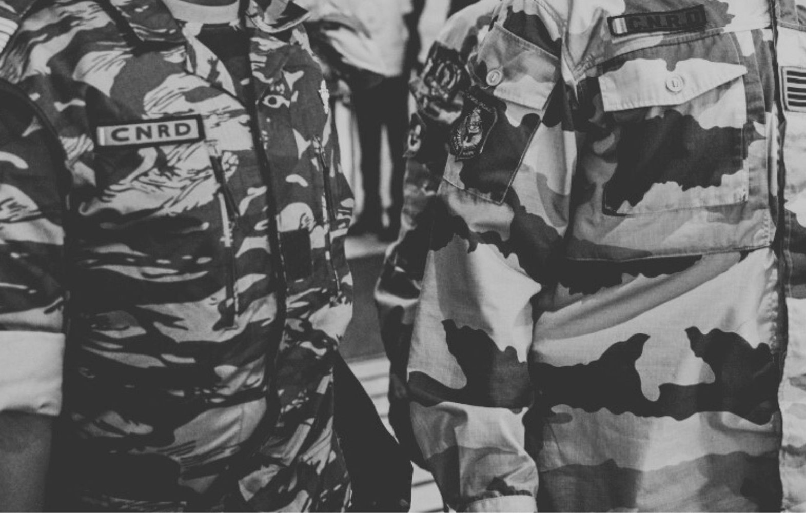 L'image montre des membres de forces armées ou de sécurité, vêtus d'uniformes. À gauche, un soldat porte un uniforme camouflage aux motifs sombres, avec des insignes sur sa poitrine. À droite, un autre soldat est vêtu d'un uniforme de camouflage plus clair, également décoré d'insignes. Les deux uniformes affichent un sens de discipline et d'autorité. L'environnement autour d'eux semble être animé, suggérant une situation dynamique ou un rassemblement. Le contraste entre les motifs des uniformes souligne leur fonction militaire.