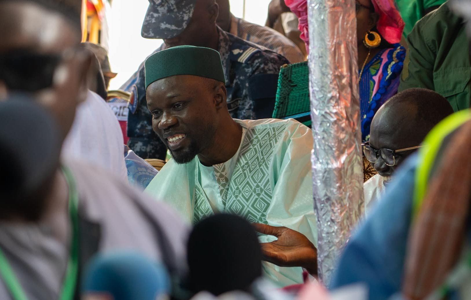 L'image montre un groupe de personnes rassemblées dans un environnement animé, probablement lors d'un événement public. Au premier plan, un homme souriant est vêtu d'un habit traditionnel, avec un turban ou une coiffe verte. Il semble engagé dans une discussion, ses mains gesticulant pour accompagner ses paroles. En arrière-plan, d'autres personnes sont présentes, certaines en uniforme, suggérant un contexte formel ou officiel. La scène est remplie de couleurs vives, et l'atmosphère semble dynamique et interactive.
