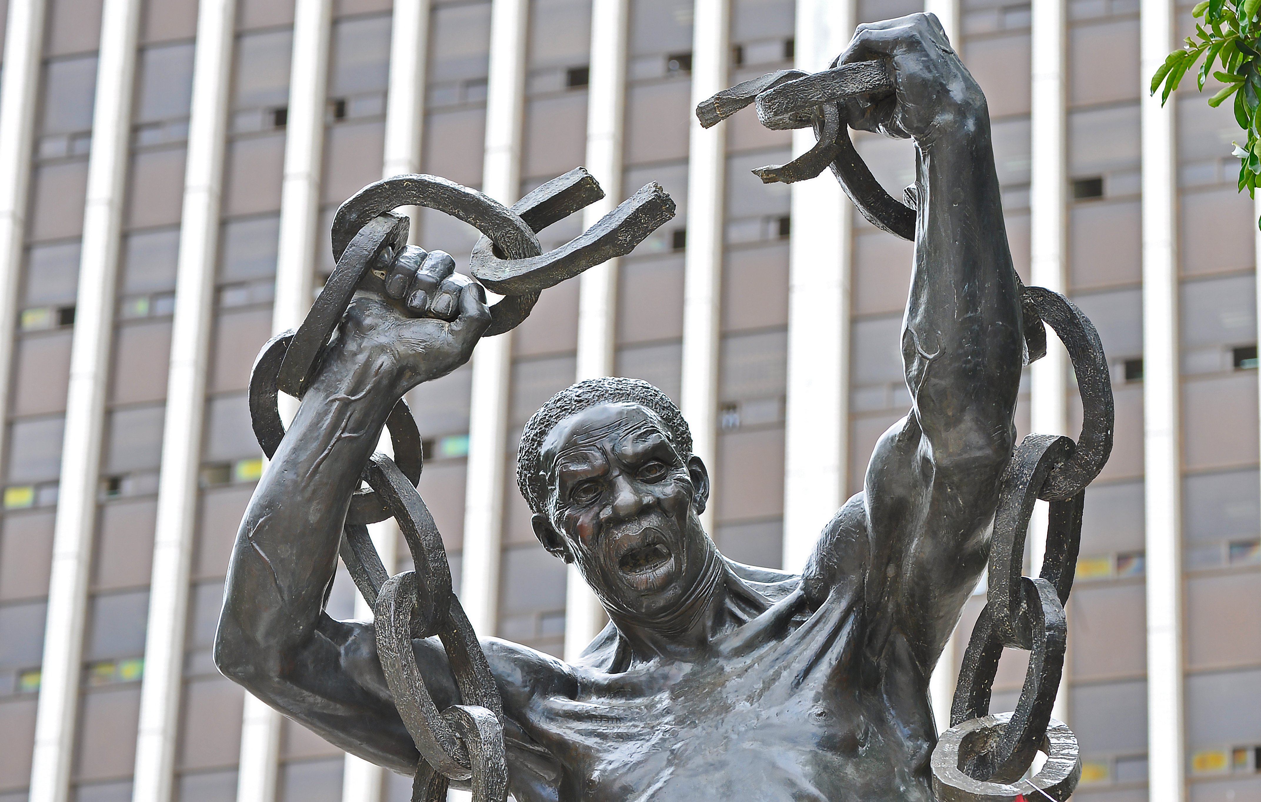 Cette image représente une statue en bronze d'un homme robuste. Il se tient debout, les bras levés au-dessus de sa tête, brandissant de lourdes chaînes brisées, symbolisant la libération et la force. Son visage exprime une intense émotion, avec une bouche ouverte comme s'il poussait un cri de victoire ou de défi. La sculpture est placée devant un bâtiment moderne, avec des lignes verticales qui soulignent contraste entre le passé et le présent. L'œuvre évoque des thèmes de résilience et de lutte pour la liberté.