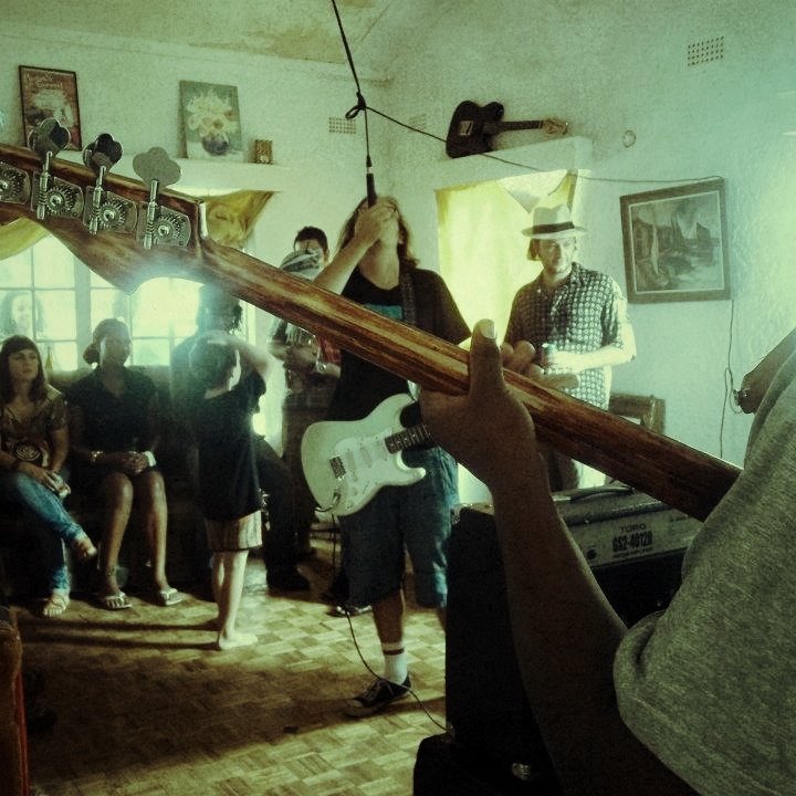 L'image montre une scène musicale vivante dans une pièce chaleureuse. À l'avant, on voit un musicien tenant une basse, tandis qu'un autre, avec une guitare électrique, se concentre sur sa performance. Les membres du groupe sont entourés d'un public qui semble apprécier la musique. Les visages sont tournés vers les musiciens, certains se balançant au rythme. La pièce est décorée avec des tableaux aux murs et des rideaux qui laissent filtrer une lumière douce. L'ambiance est conviviale, remplie d'énergie et de passion pour la musique.