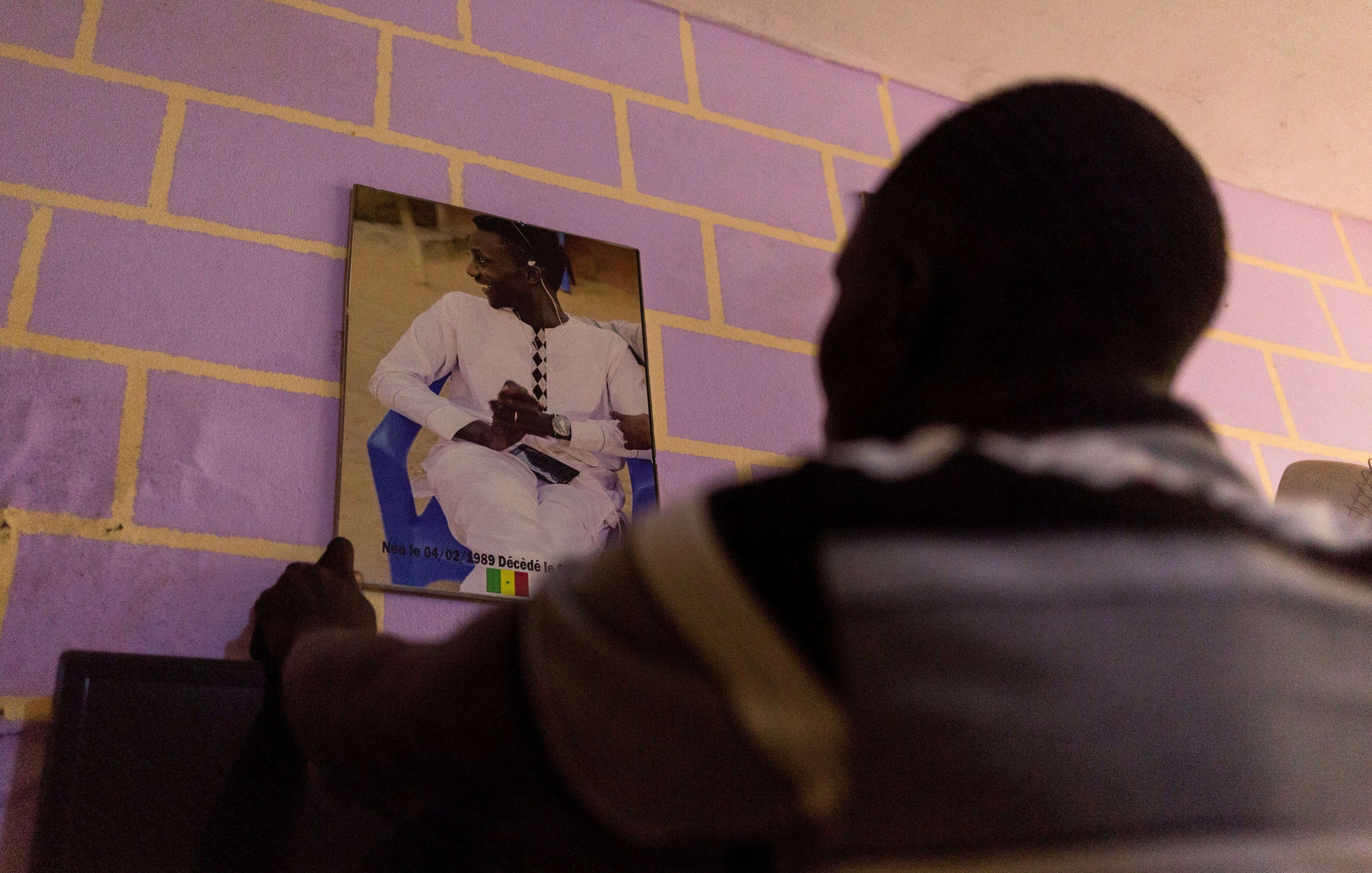 Dans cette image, un homme est en train de fixer un portrait au mur. Il se tient de dos, face à un mur peint en violet aux briques jaunes. Le portrait montre une personne assise, vêtue d'une tenue traditionnelle, probablement sénégalaise, avec un fond flou. L'homme qui fixe le portrait semble concentré sur sa tâche, ajoutant une touche personnelle à l'espace où il se trouve.