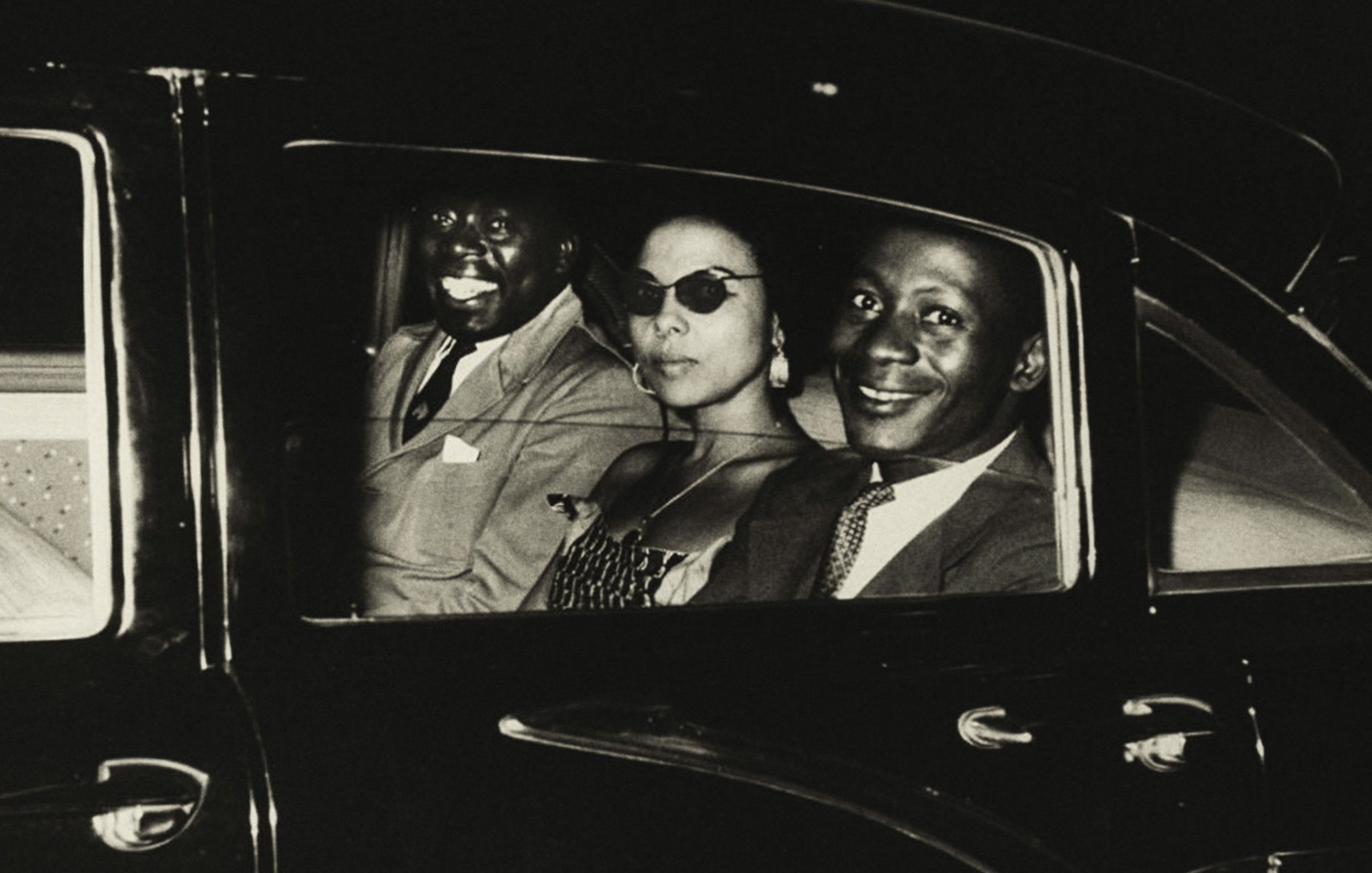 L'image présente trois personnes assises à l'intérieur d'une voiture sombre. L'ambiance semble élégante et vintage. À gauche, un homme souriant, au teint foncé, porte un costume clair. Au centre, une femme avec des lunettes de soleil stylées, habillée d'une robe à motifs, dégage une aura de sophistication. À droite, un autre homme, également souriant, avec un teint légèrement plus clair, porte une tenue qui semble chic. Les visages des occupants sont mis en valeur par un éclairage doux, créant une atmosphère chaleureuse et amicale. Le tout suggère un moment de convivialité, peut-être après une sortie ou un événement.