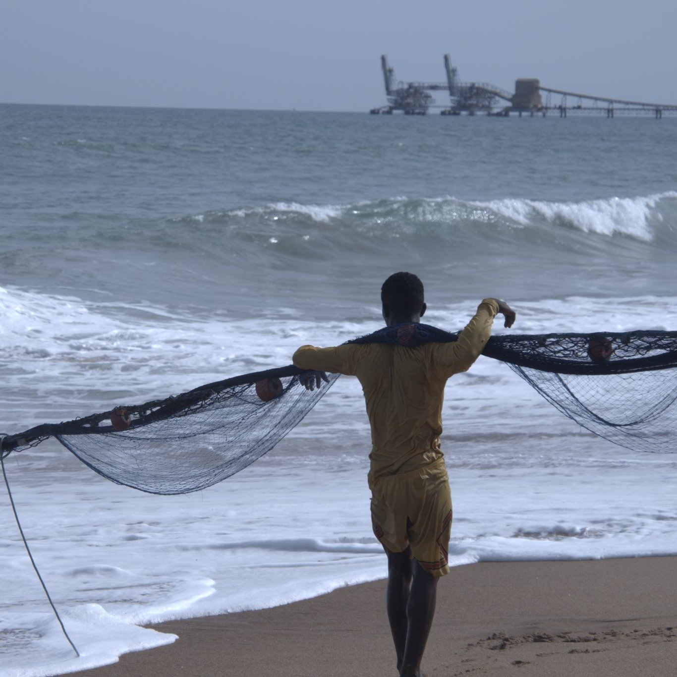 Sur cette image, un homme se trouve sur une plage, dos à nous. Il porte un maillot de bain jaune et tire un grand filet de pêche, qui s'étend devant lui. Les vagues de l'océan s'écrasent doucement sur le sable, produisant un bruit apaisant. À l'arrière-plan, on aperçoit une jetée où se trouvent des structures industrielles, ajoutant une touche de la vie maritime. Le ciel est clair et lumineux, créant une ambiance de tranquillité et de travail en harmonie avec la mer.