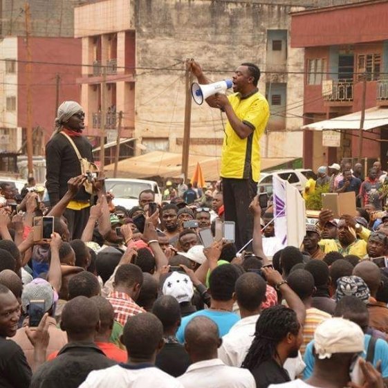 L'image montre une scène de rassemblement dans une rue animée, remplie de nombreuses personnes. Au centre, un orateur, habillé en jaune, se tient sur une plateforme surélevée, tenant un mégaphone et s'adressant à la foule. Les membres de l'audience, qui semblent nombreux et engagés, lèvent souvent leurs téléphones, capturant probablement des moments du discours. En arrière-plan, on aperçoit des bâtiments et une affichage indiquant des prix, ce qui suggère un environnement urbain. L'atmosphère est énergique, marquée par l'enthousiasme et la mobilisation des participants.
