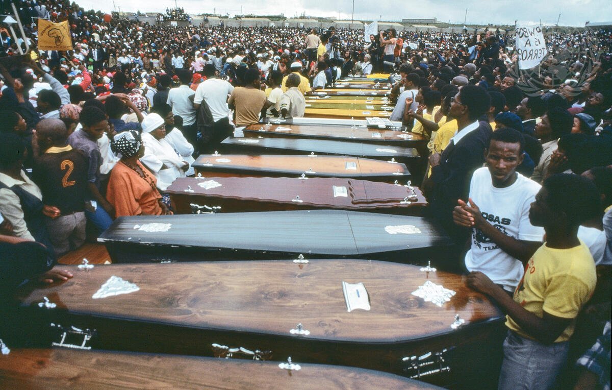 L'image montre une vaste foule rassemblée en extérieur, entourant une série de cercueils alignés. Les cercueils, de différentes couleurs et styles, sont disposés côte à côte, symbolisant une cérémonie funèbre. Les personnes présentes semblent nombreuses et engagées, certaines portent des vêtements de deuil, tandis que d'autres affichent des expressions de tristesse ou de solidarité. L'atmosphère est empreinte de respect et de recueillement, évoquant une perte collective. Au fond, des pancartes sont visibles, ajoutant une dimension politique ou sociale à l'événement. Le ciel est nuageux, renforçant le caractère solennel de la scène.