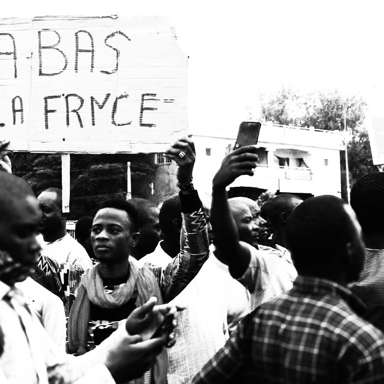 L'image représente une scène de manifestation. Au premier plan, plusieurs personnes se rassemblent, certaines levant les bras. Un homme, au centre, tient une grande pancarte avec l'inscription "ABAS LA FRYCE" en lettres capitales. La foule semble engagée et énergique, exprimant une certaine frustration ou un message de contestation. L'ambiance est chargée d'émotion, avec des visages déterminés et des gestes expressifs, dans un cadre urbain qui suggère un lieu public. Les vêtements des manifestants varient, ajoutant à la diversité de la scène.