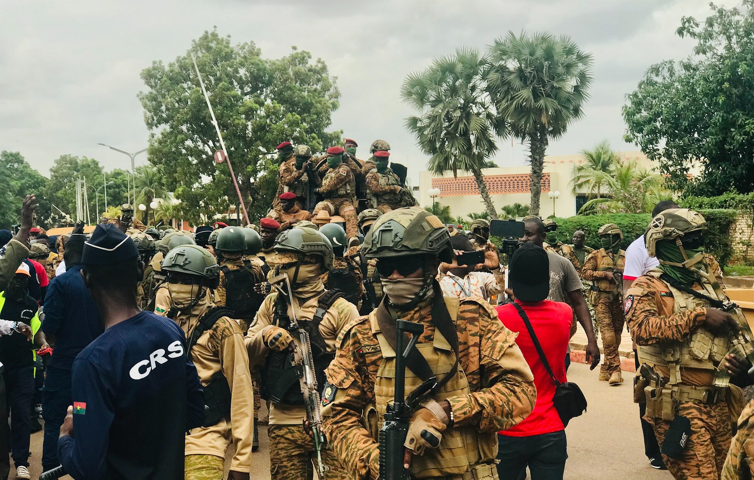The image depicts a scene of armed soldiers in a public area. The soldiers are wearing camouflage uniforms and helmets, some with face coverings, and are equipped with rifles. In the background, there are additional soldiers on a vehicle. The atmosphere appears to be tense, with a gathering of civilians present as well, some taking photographs. There are trees and buildings in the background, suggesting an urban setting. Overall, the scene conveys a strong military presence.