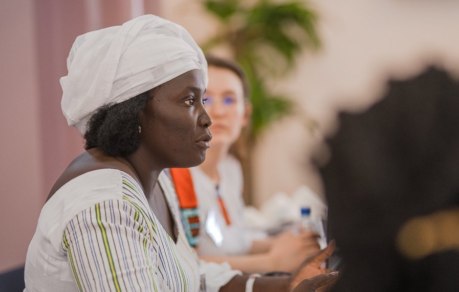 L'image montre une femme assise à une table lors d'une réunion ou d'une discussion. Elle porte une tenue traditionnelle, avec une chemise à rayures et un foulard blanc sur la tête. Son expression est sérieuse et concentrée, et elle semble engagée dans la conversation. À côté d'elle, une autre femme est présente, mais elle est floue en arrière-plan. On peut apercevoir des bouteilles d'eau sur la table, ainsi qu'une ambiance chaleureuse et collaborative. Des éléments de verdure, comme une plante, ajoutent une touche de couleur à la scène.