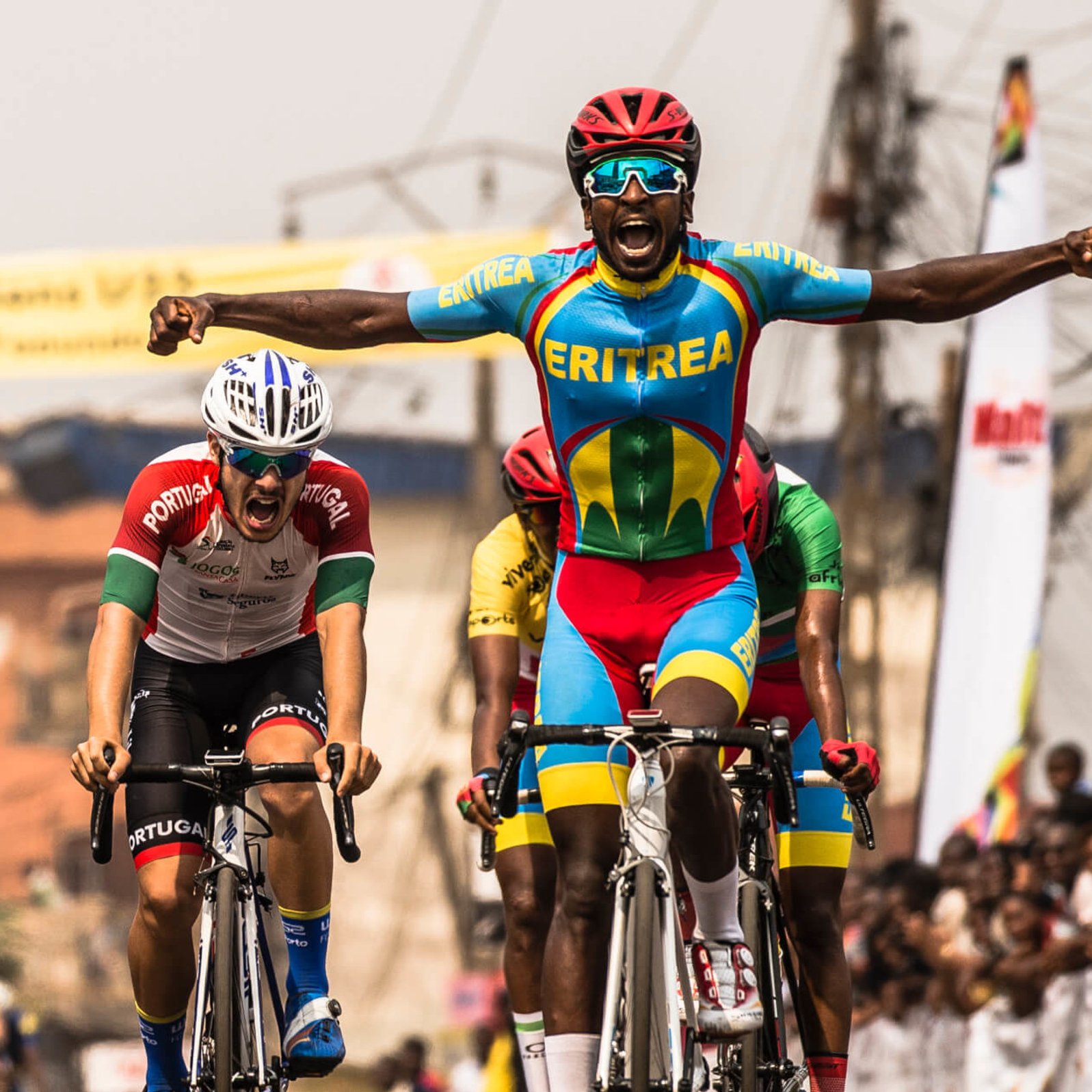 L'image montre un moment dynamique d'une course de cyclisme. Au premier plan, un coureur, vêtu d'un maillot multicolore représentant l'Érythrée, lève les bras en signe de victoire, affichant une expression de joie et d'excitation. En arrière-plan, un autre cycliste, portant un maillot aux couleurs du Portugal, semble montrer des signes d'effort intensif, presque à la limite de l'épuisement. On aperçoit également d'autres participants et une foule enjouée de spectateurs, créant une ambiance festive autour de cet événement sportif. Le décor est vibrant, évoquant l'énergie et la compétitivité de la course.