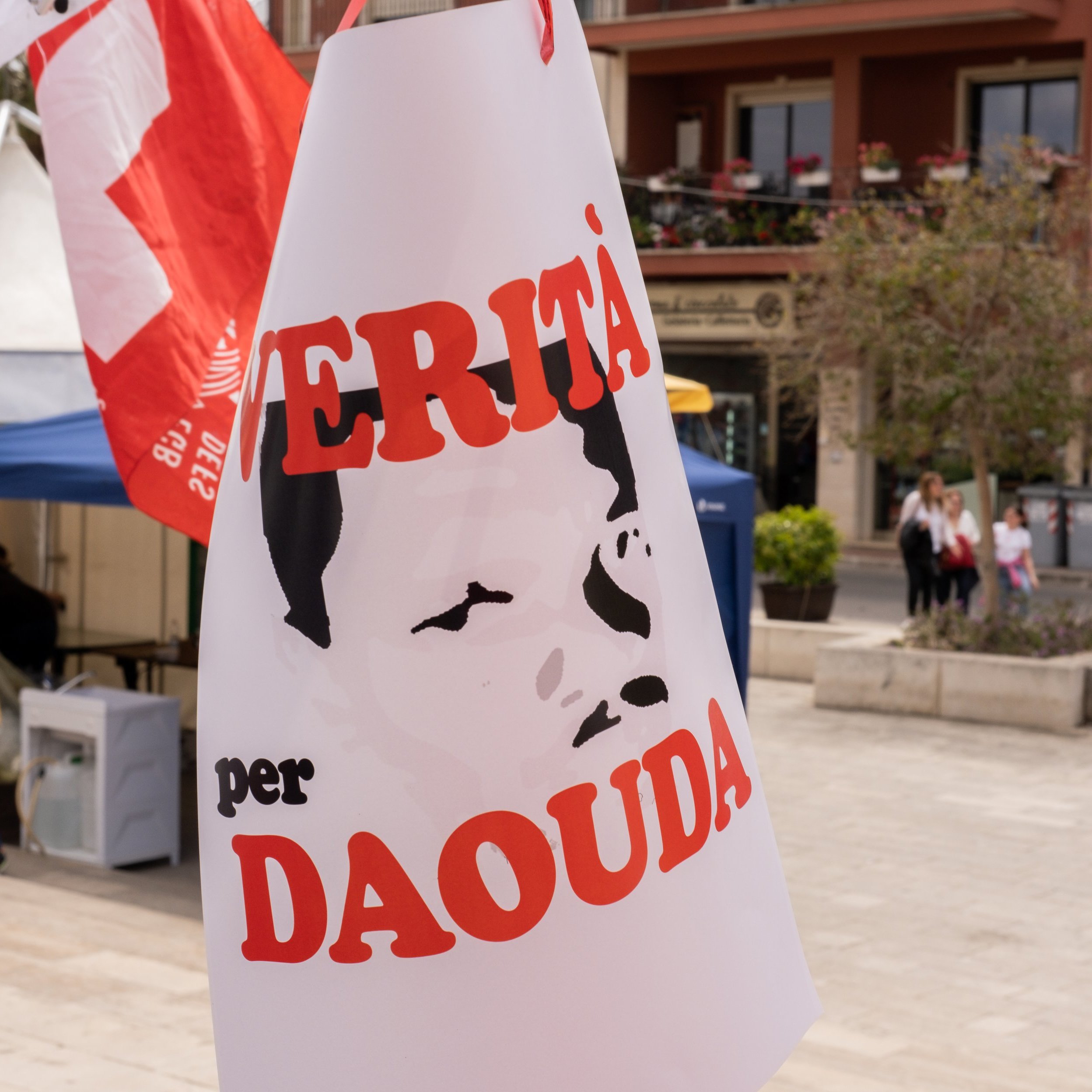 L'image présente une scène en plein air, probablement lors d'un événement ou d'une manifestation. Un panneau est palpable, affichant un portrait stéréotypé d'une personne, dont les traits sont simplifiés et en noir et blanc. Le texte en rouge indique "VERITÀ per DAOUDA", ce qui signifie "Vérité pour Daouda". En arrière-plan, on aperçoit des bâtiments avec des balcons et des tentes blanches. L'atmosphère semble engagée, avec un mélange de couleurs vives des drapeaux et des vêtements des personnes présentes. Le son des conversations et des rires pourrait également emplir l'air, créant une ambiance dynamique et communautaire.
