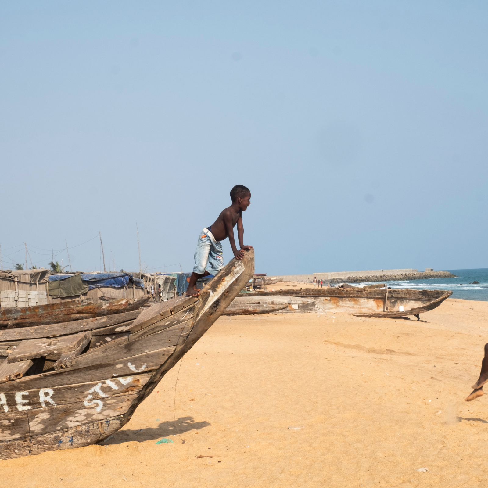 Dans cette scène vibrante, une femme porte une grande bassine sur sa tête, symbole de son travail quotidien. Elle avance avec assurance sur un chemin, tout en étant entourée de maisons à architecture simple, en bois et en bambou. Le ciel est clair, laissant présager une belle journée ensoleillée. À proximité, des enfants jouent ensemble, ajoutant une atmosphère de vie et de dynamisme à l'environnement. Les ombres et les textures des matériaux environnants se mêlent, créant un paysage riche et vivant. L'ensemble dégage une ambiance d'activité et de communauté.