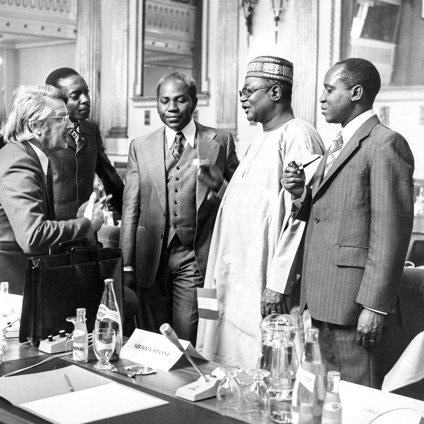 L'image représente un groupe de cinq hommes en discussion dans un cadre formel, probablement lors d'une conférence ou d'une réunion. Ils sont habillés en costumes élégants, dont certains sont accompagnés de cravates. Les visages montrent une interaction animée, avec des gestes de mains qui suggèrent une conversation engageante. En arrière-plan, on aperçoit des drapeaux, ce qui indique un contexte international. La scène se déroule dans une grande salle, ornée et bien éclairée, ajoutant une atmosphère de sérieux et de respect.