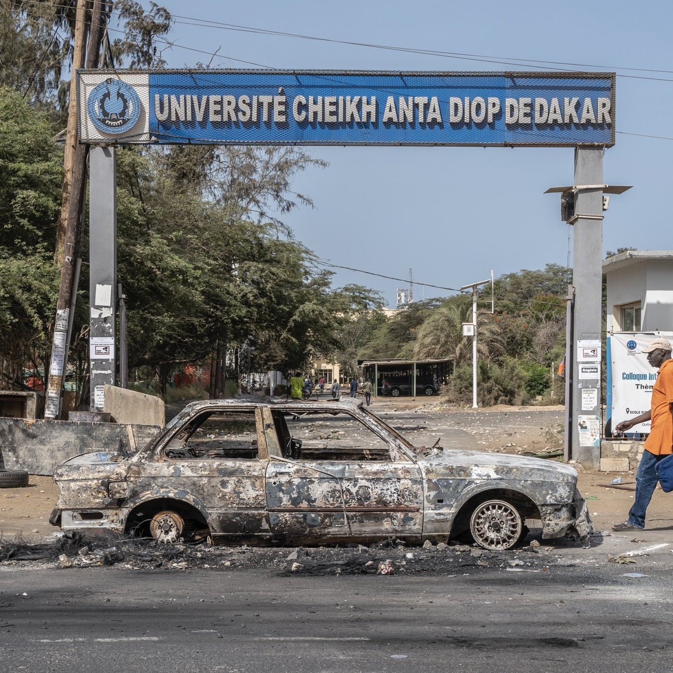 L'image montre l'entrée de l'Université Cheikh Anta Diop de Dakar, identifiable par le panneau au-dessus. En premier plan, une voiture calcinée est garée sur la route, avec des traces de brûlures et des dégâts visibles, indiquant qu'elle a été abandonnée après un incendie. En arrière-plan, on peut apercevoir des arbres et des structures, mais l'atmosphère semble marquée par un sentiment de désolation. Un piéton, portant une sacoche, marche sur le trottoir, ajoutant un contraste entre l'activité humaine et l'état dégradé des lieux.