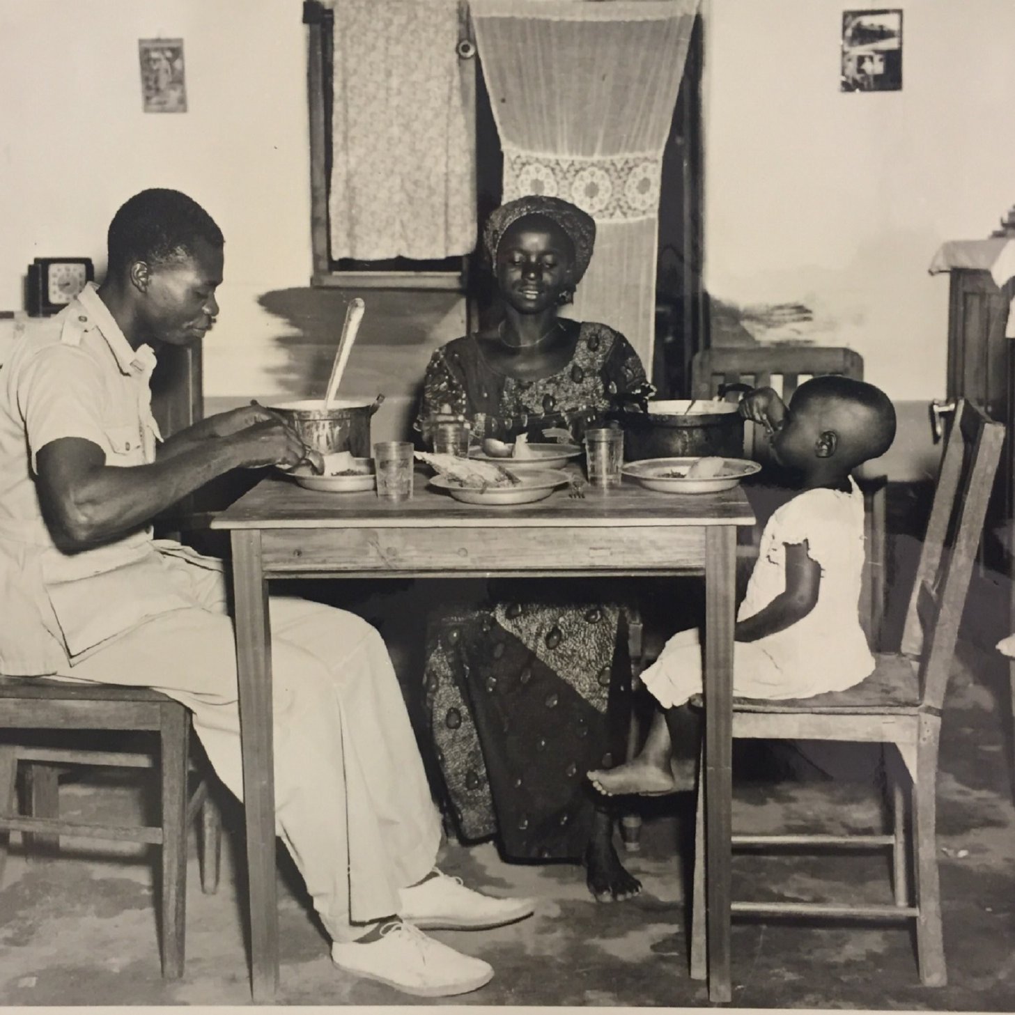 L'image en noir et blanc montre une scène de repas familial dans une pièce simple, probablement une cuisine. À une table en bois, un homme en uniforme, assis sur une chaise, semble être en train de manger avec une grande cuillère. En face de lui, une femme, vêtue d'une robe traditionnelle et d'un chapeau, regarde avec un sourire bienveillant. À côté d'elle, un petit enfant est assis sur une autre chaise, portant une petite robe blanche, observant la scène. Les murs de la pièce sont décorés sobrement, et il y a des verres sur la table, ajoutant une touche de quotidien à ce moment plein de convivialité.