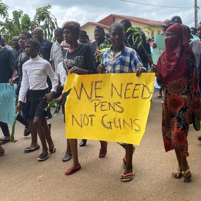 Dans cette image, un groupe de personnes marche ensemble dans une rue. On peut entendre des voix et des chants de solidarité. Les participants tiennent des pancartes aux couleurs vives. Certaines pancartes affichent des messages puissants comme "WE NEED PENS NOT GUNS", exprimant une demande pour la paix et l'éducation plutôt que pour la violence. Les manifestants sont vêtus de différents styles de vêtements, allant de tenues formelles à des vêtements décontractés. En arrière-plan, on aperçoit des maisons et des arbres qui ajoutent un cadre naturel à cette scène de protestation pacifique. L'atmosphère est déterminée et engagée, reflet d'un désir de changement et d'un message fort pour la communauté.