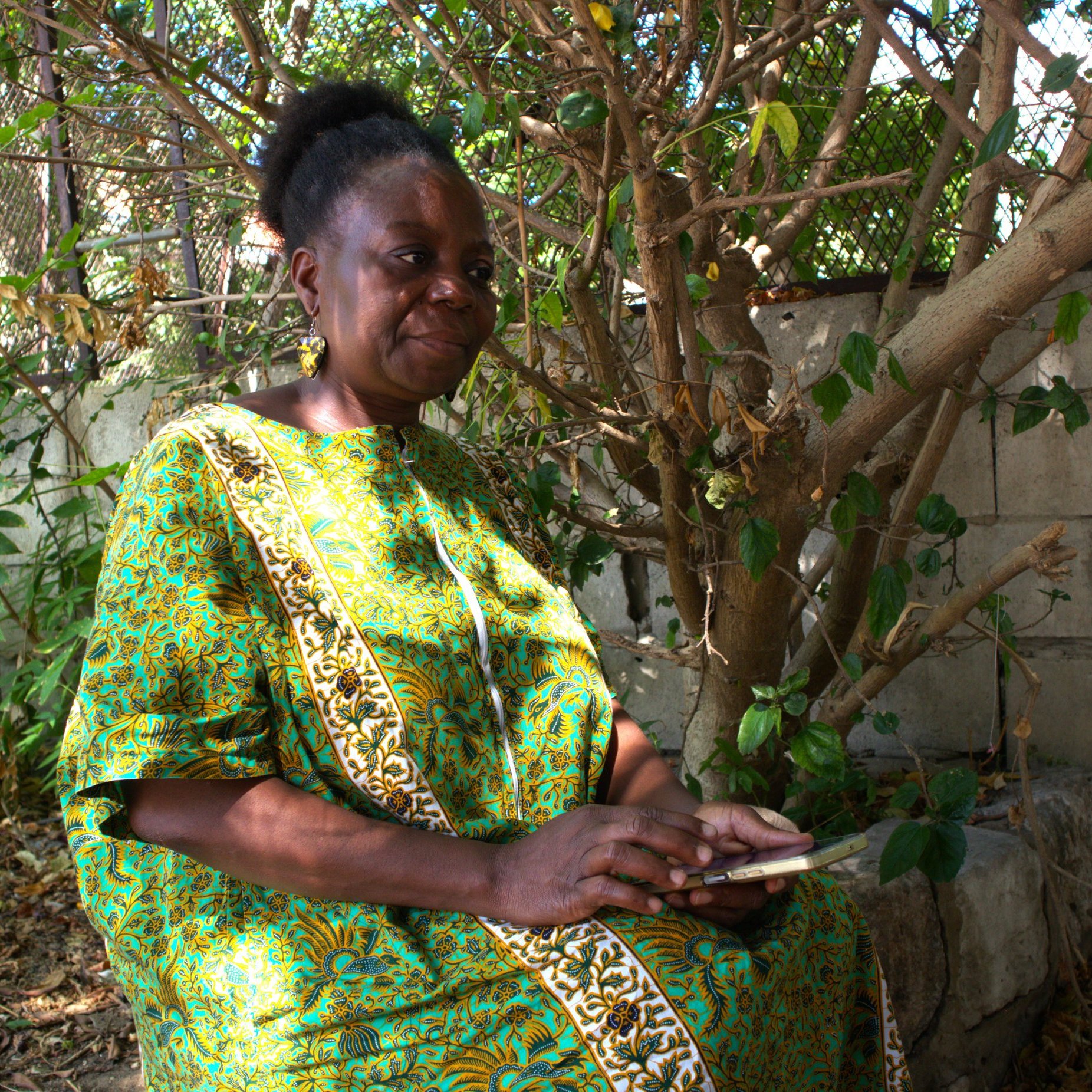 Dans cette image, une femme est assise sous un arbre. Elle porte une robe traditionnelle colorée, dominée par des nuances de vert et des motifs vibrants. Son visage exprime une sérénité et une douce réflexion. À côté d'elle, on peut voir une surface en pierre qui pourrait être un mur. La lumière naturelle filtre à travers les feuillages de l'arbre, créant une atmosphère paisible et intime. Il semble qu'elle tient un objet dans ses mains, probablement un téléphone, et elle est entourée de verdure, ce qui ajoute une touche de vie à la scène.