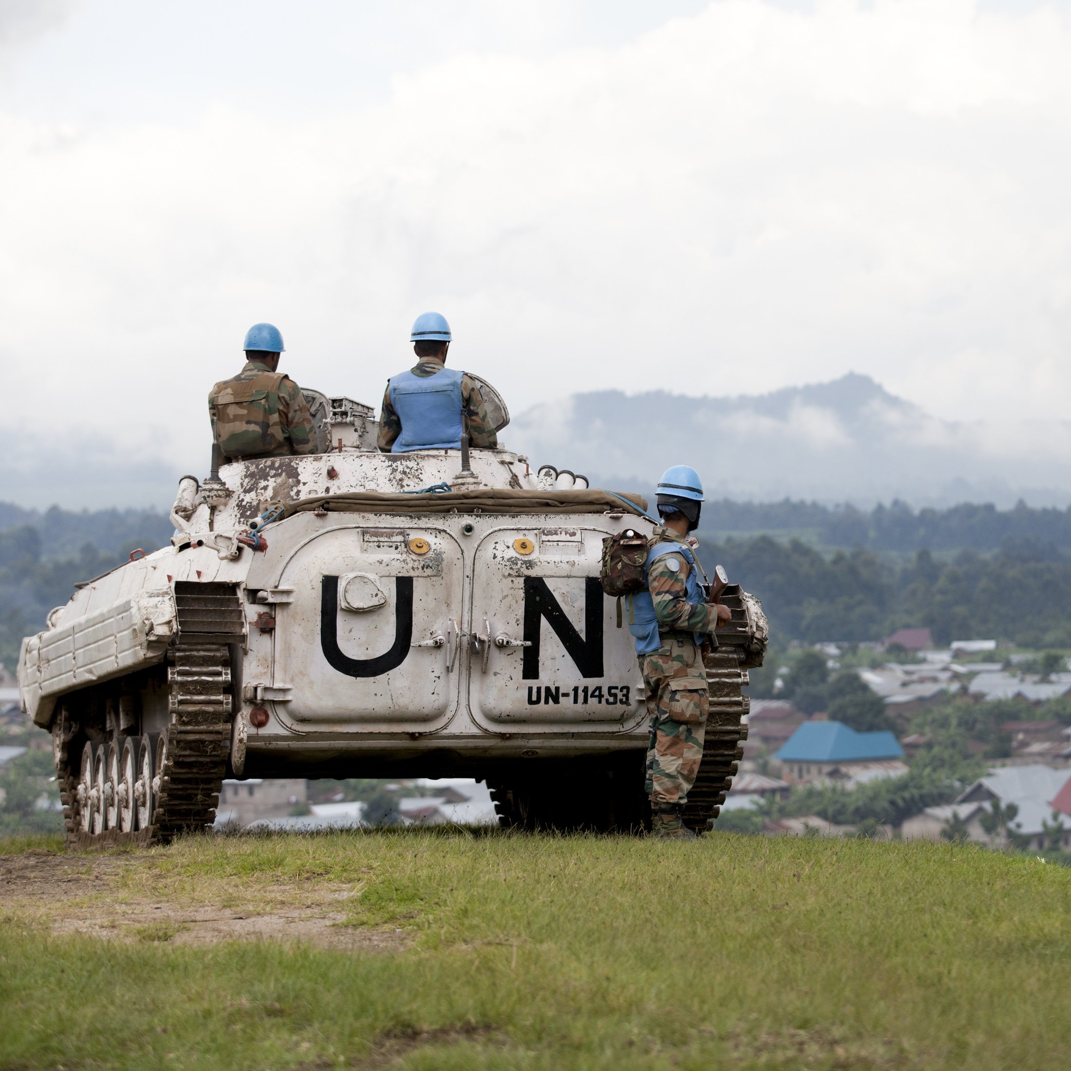 L'image présente un paysage où des soldats en uniforme de l'ONU se trouvent autour d'un véhicule blindé. Le véhicule, marqué par le symbole "UN" en lettres blanches, est stationné sur une colline qui surplombe une petite ville. Les soldats portent des casques bleu clair typiques des missions de maintien de la paix. À l'arrière-plan, on aperçoit des maisons aux toits colorés et une végétation dense, tandis que le ciel est partiellement nuageux, créant une atmosphère sereine malgré le contexte militaire. Les soldats semblent vigilants et concentrés, surveillant les environs.