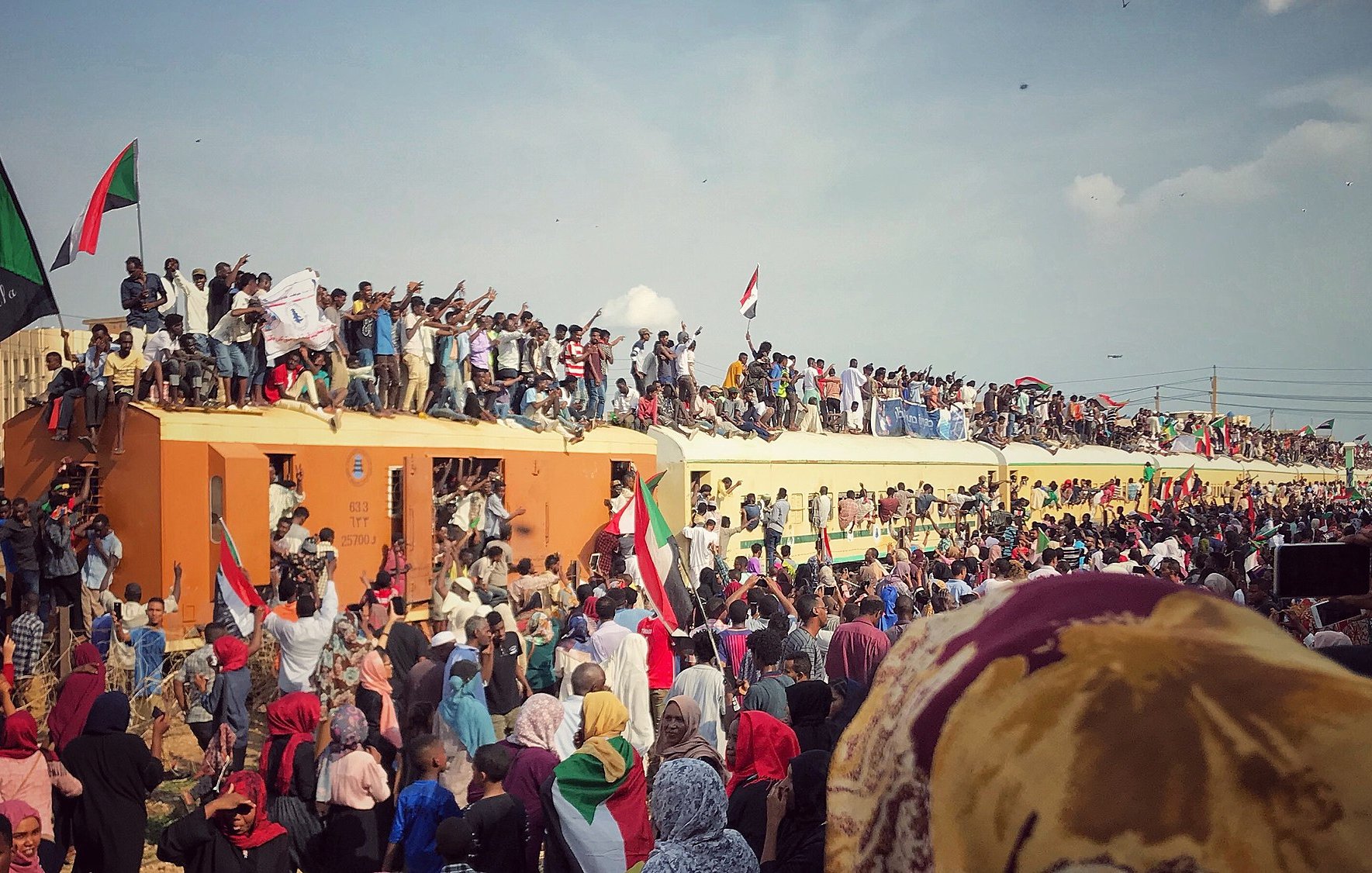L'image montre une scène de grande effervescence, probablement lors d'un rassemblement ou d'une célébration. On peut voir une foule nombreuse, avec des gens debout sur un train, brandissant des drapeaux aux couleurs vives, en particulier du rouge, du noir et du vert. Les participants expriment leur enthousiasme et leur solidarité, certains levant les poings ou affichant des panneaux. La foule est diverse, incluant des personnes de tous âges, et il règne une atmosphère de ferveur collective, avec un ciel bleu parsemé de quelques nuages en arrière-plan. Cette image évoque des sentiments de joie, d'unité et de détermination.