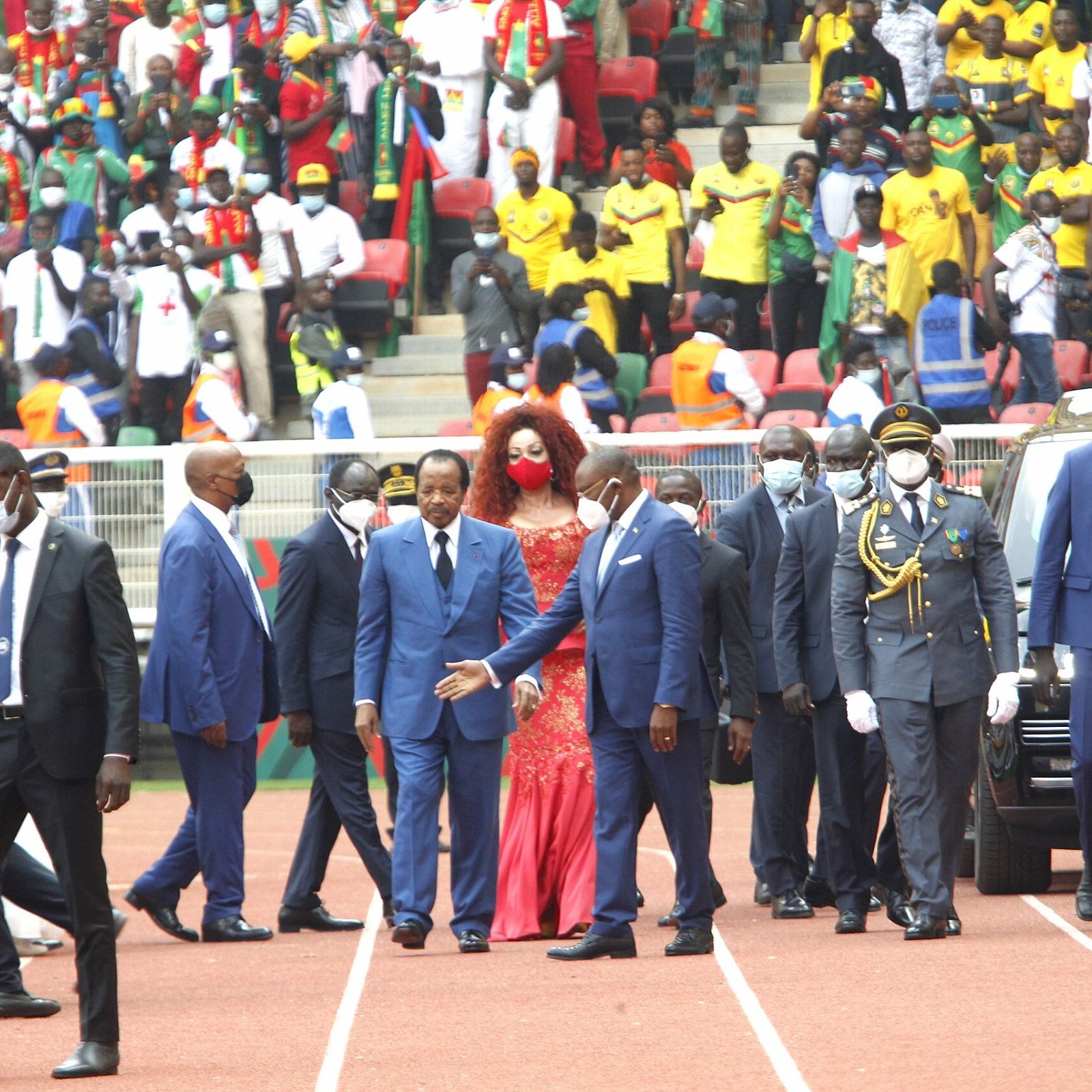 L'image montre un événement en extérieur, probablement un stade rempli de spectateurs. Au premier plan, un groupe de personnalités, dont plusieurs hommes en costume sombre et cravate, marche avec assurance. Parmi eux, une femme se distingue grâce à sa robe rouge éclatante et sa coiffure volumineuse. Ils semblent être en route pour un moment important, entourés de sécurité. À l'arrière-plan, on peut deviner une foule colorée et animée, avec des supporters portant divers vêtements aux couleurs vives, témoignant d'une atmosphère festive. L'ambiance générale semble être celle d'une grande célébration ou d'une cérémonie officielle.