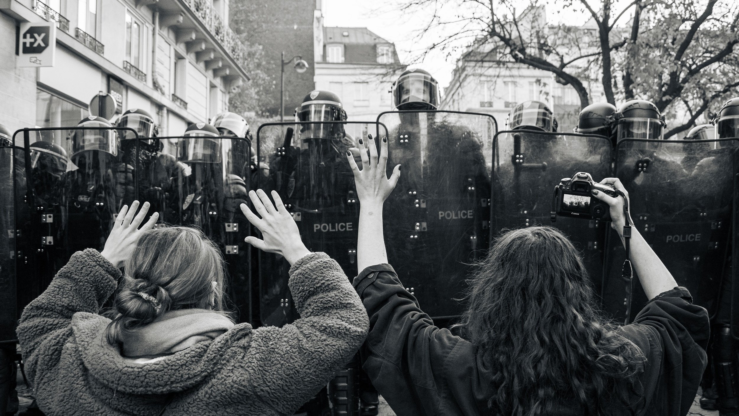 L'image montre une scène de manifestation en noir et blanc. À l'avant-plan, deux personnes se tiennent face à des policiers en tenue anti-émeute. Les manifestants, une femme brune avec les cheveux longs et une autre avec des cheveux bouclés, lèvent les bras en signe de protestation ou de désescalade. Les policiers sont rangés en formation derrière des boucliers, créant une barrière. L'environnement urbain est visible en arrière-plan, avec des bâtiments peut-être typiques de la ville. L'atmosphère est tendue, reflétant un moment de confrontation pacifique mais chargée d'émotion.