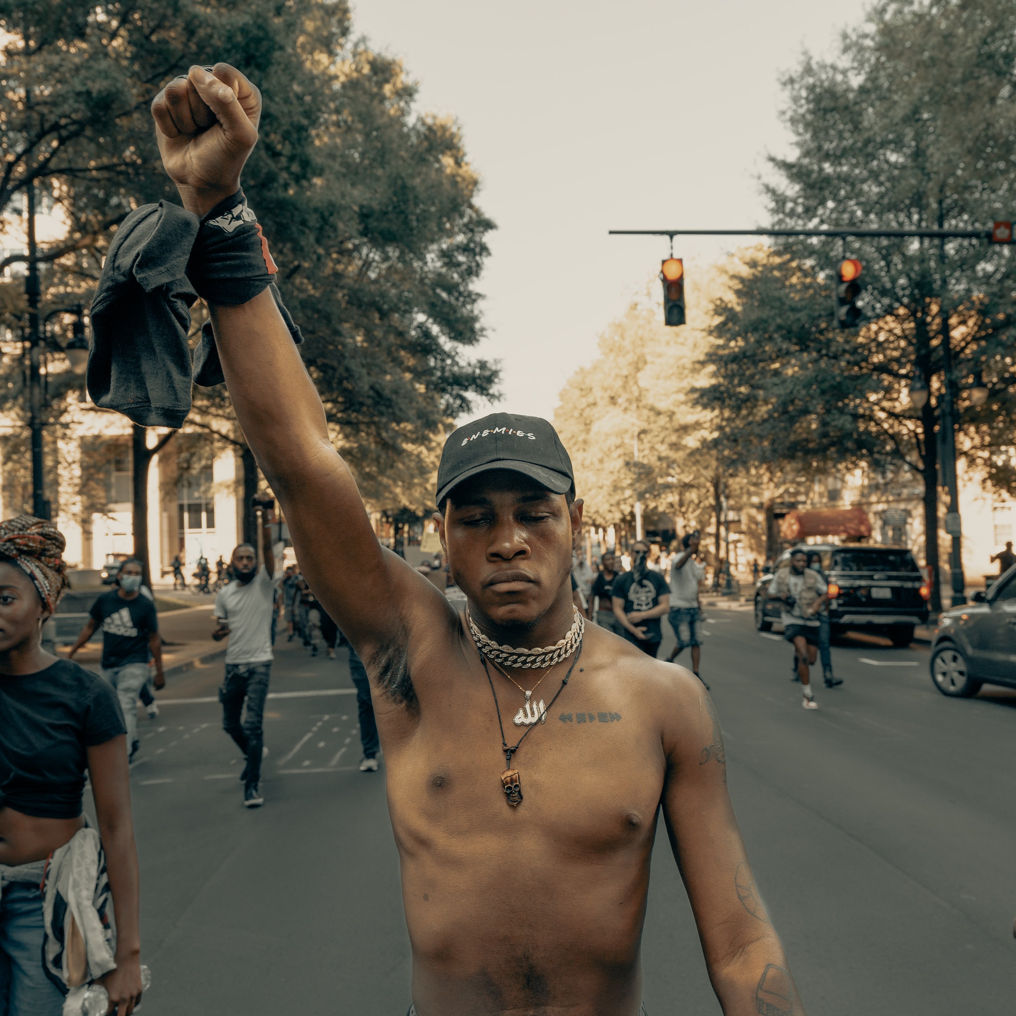L'image montre une scène de manifestation dans une rue. Au premier plan, un jeune homme, torse nu, lève le poing en signe de protestation. Il porte une casquette et des chaînes autour du cou. À l'arrière-plan, d'autres personnes marchent, certaines levant également le poing, témoignant d'un esprit collectif. La rue est bordée d'arbres, et on peut apercevoir des voitures et des feux de signalisation. L'ambiance semble énergique et engagée, reflétant une lutte pour la justice ou l'égalité.