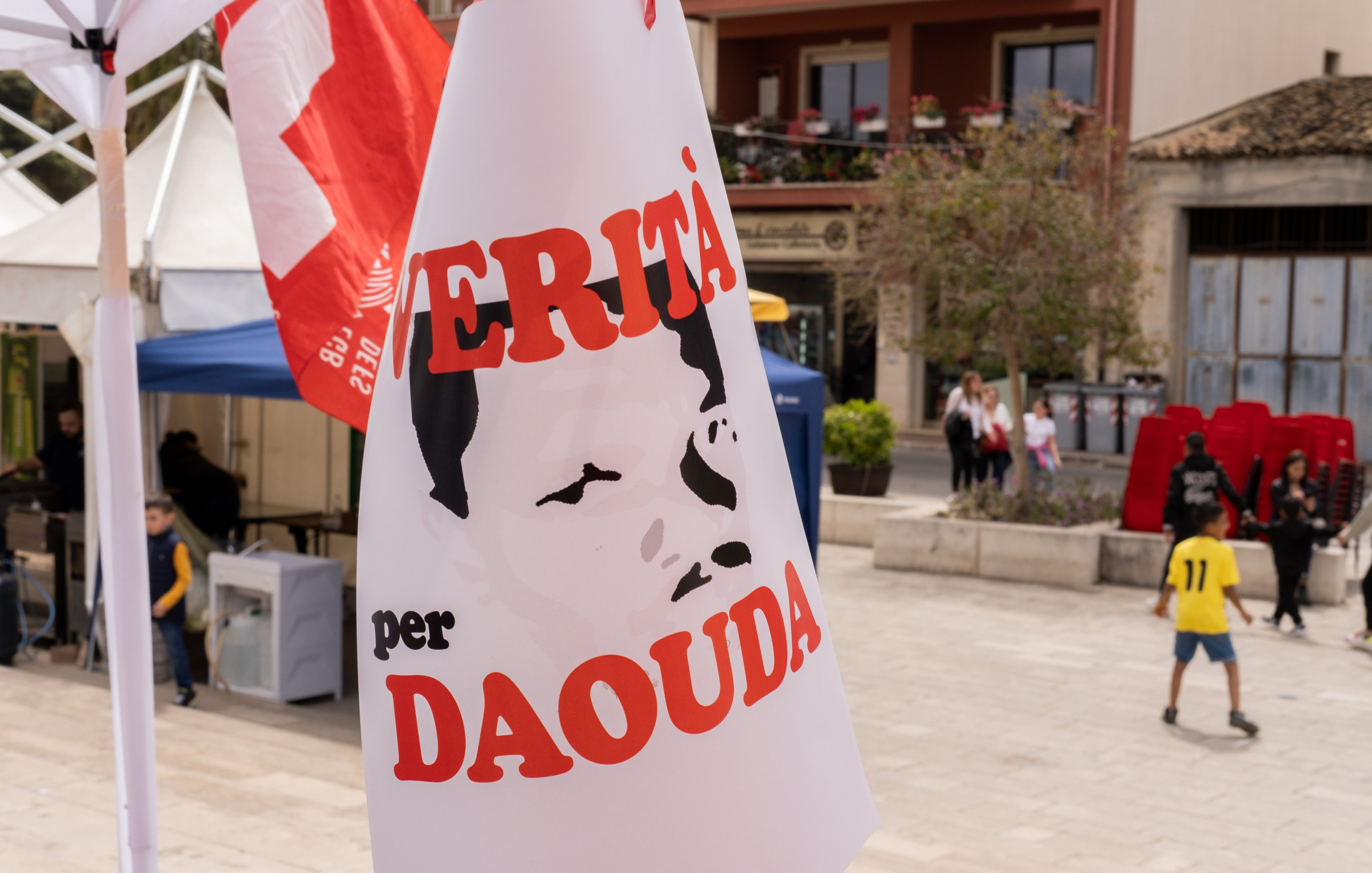 L'image présente une scène en plein air, probablement lors d'un événement ou d'une manifestation. Un panneau est palpable, affichant un portrait stéréotypé d'une personne, dont les traits sont simplifiés et en noir et blanc. Le texte en rouge indique "VERITÀ per DAOUDA", ce qui signifie "Vérité pour Daouda". En arrière-plan, on aperçoit des bâtiments avec des balcons et des tentes blanches. L'atmosphère semble engagée, avec un mélange de couleurs vives des drapeaux et des vêtements des personnes présentes. Le son des conversations et des rires pourrait également emplir l'air, créant une ambiance dynamique et communautaire.