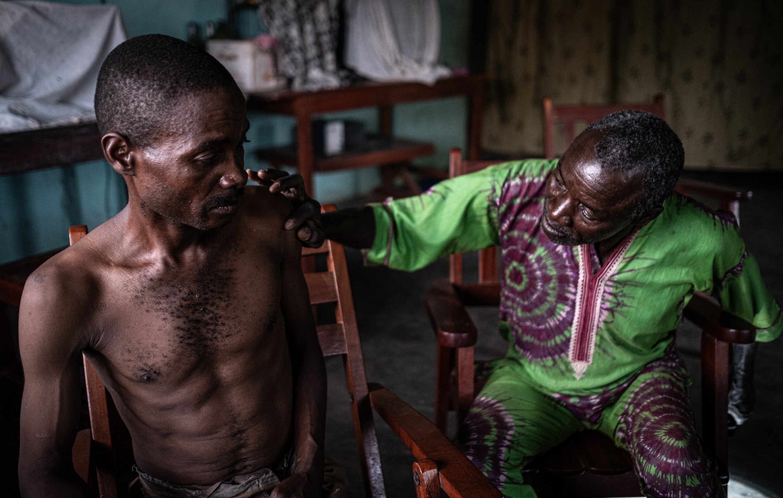 L'image montre deux hommes assis dans des chaises en bois dans une pièce intérieure. L'un des hommes, assis à gauche, a un torse nu et semble pensif. Son expression montre une certaine vulnérabilité. L'autre homme, à droite, est vêtu d'une robe de couleur verte et violette, et il semble s'adresser au premier avec attention, posant une main réconfortante sur son épaule. La lumière dans la pièce est douce, créant une atmosphère intimiste. En arrière-plan, on peut voir des objets flous disposés sur des tables, ce qui suggère un environnement familier et intime.