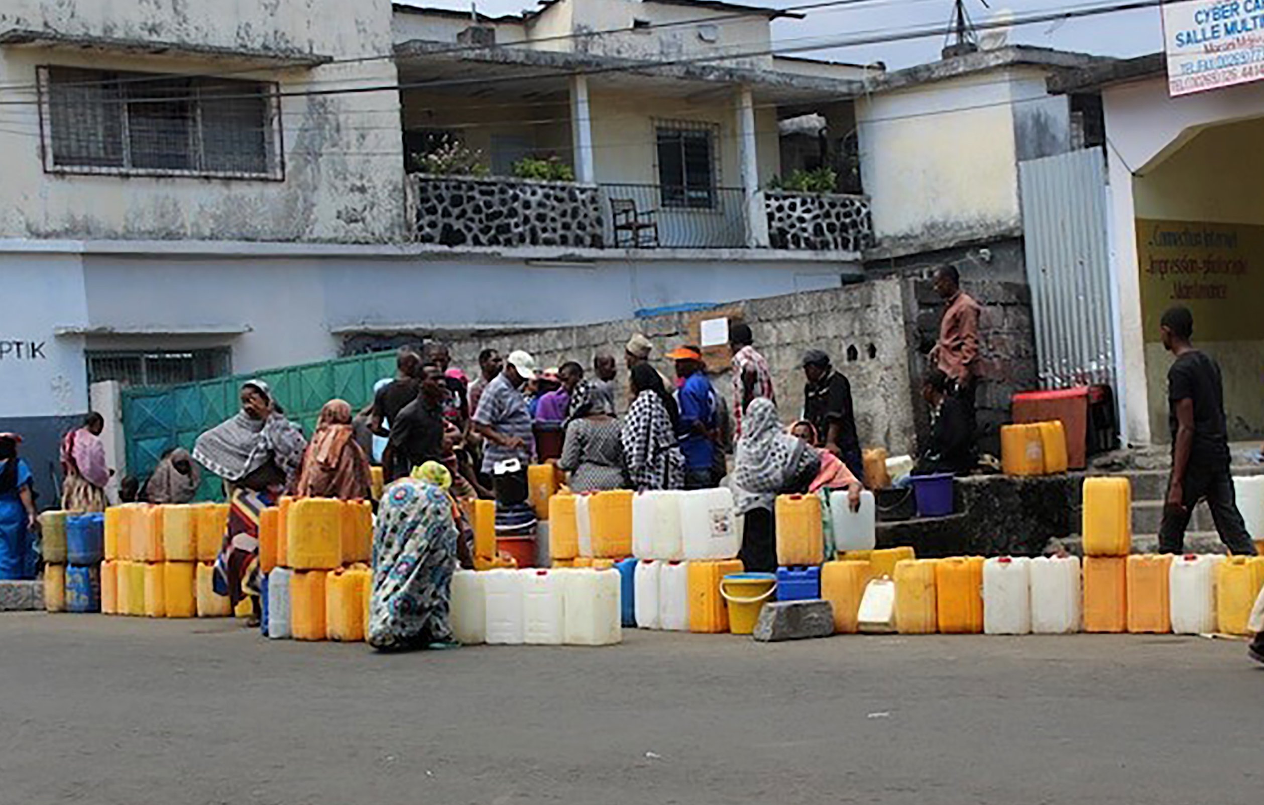 L'image montre une scène animée dans une rue. Des groupes de personnes se rassemblent autour de plusieurs conteneurs en plastique de différentes tailles, souvent de couleur orange ou jaune. Ces conteneurs sont alignés sur le sol. Les personnes, principalement habillées de vêtements traditionnels, semblent attendre leur tour pour remplir les récipients d'eau. En arrière-plan, on distingue des bâtiments aux façades simples, certains avec des balcons. L'ambiance est celle d'une activité quotidienne, soulignant l'importance de l'eau dans la vie des gens.