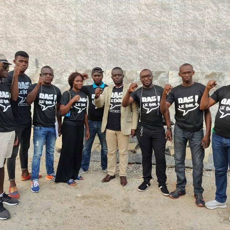 L'image montre un groupe de onze personnes rassemblées, certaines portant des t-shirts noirs avec des inscriptions. Ils affichent tous des expressions déterminées et se tiennent debout, avec quelques-uns levant le poing en signe de solidarité ou de protestation. Les membres du groupe sont de différents sexes et portent des vêtements variés, allant de t-shirts décontractés à des vêtements plus formels. L'arrière-plan est constitué d'un mur en pierre, ajoutant une texture neutre à la scène. L'ambiance semble énergique et engagée.