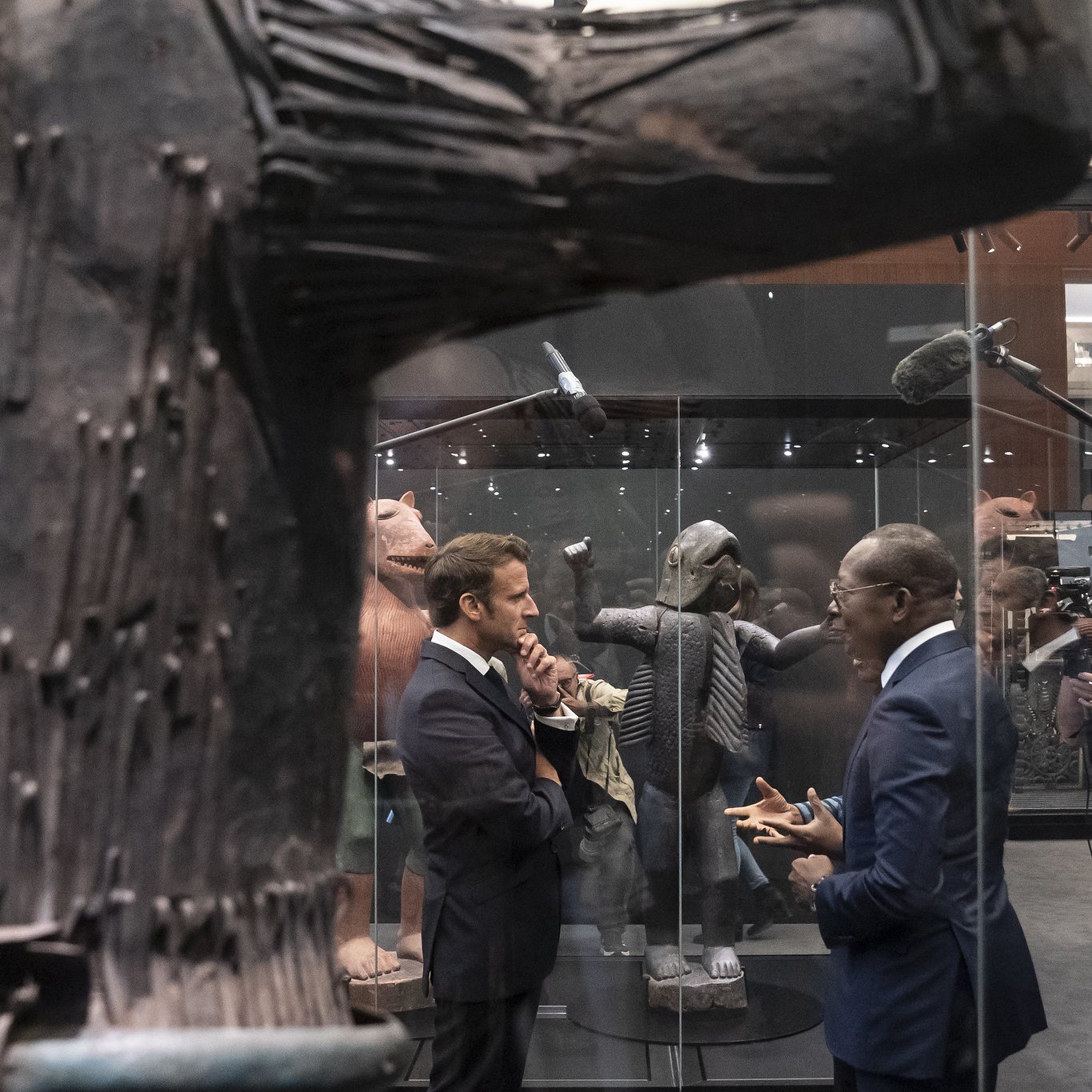 L'image montre un espace d'exposition où deux hommes en costume discutent. L'un d'eux semble concentré sur la conversation, tandis que l'autre écoute attentivement. Ils se tiennent près d'une grande sculpture en bois, qui occupe une place centrale. Cette sculpture a une texture rugueuse, avec des détails qui évoquent une forme humaine ou abstraite. En arrière-plan, d'autres œuvres d'art sont visibles, ajoutant à l'atmosphère culturelle de l'exposition. L'éclairage est doux, mettant en valeur les sculptures tout en créant une ambiance contemplative et respectueuse.