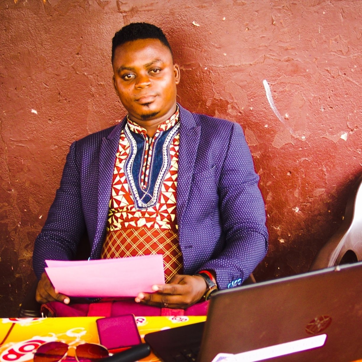 Dans l'image, un homme est assis à une table en bois, près d'un ordinateur portable noir. Il porte une chemise traditionnelle aux motifs colorés, mêlant des nuances de rouge, bleu et blanc, et une veste sombre qui contraste avec sa tenue. Son expression est calme et confiante. Sur la table, on peut voir des feuilles de papier rose ainsi que quelques objets, probablement des lunettes de soleil. Le fond est d'un brun uni, ce qui met en valeur le personnage au premier plan. L'atmosphère semble détendue et conviviale.