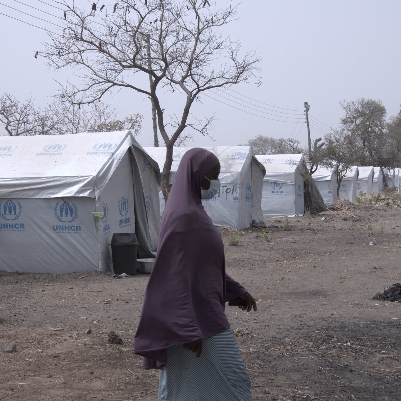 L'image montre un camp de réfugiés, caractérisé par plusieurs tentes en toile blanches et bleues, portant le logo de l'UNHCR. Au premier plan, une femme marche, vêtue d'un voile mauve et d'une robe claire, se dirigeant vers l'une des tentes. Le paysage environnant est aride et désolé, avec quelques arbres dénudés, suggérant un environnement difficile. En arrière-plan, des lignes électriques sont visibles, ce qui indique une certaine infrastructure. L'atmosphère semble calme, mais il y a une sensation de précarité et de résilience.