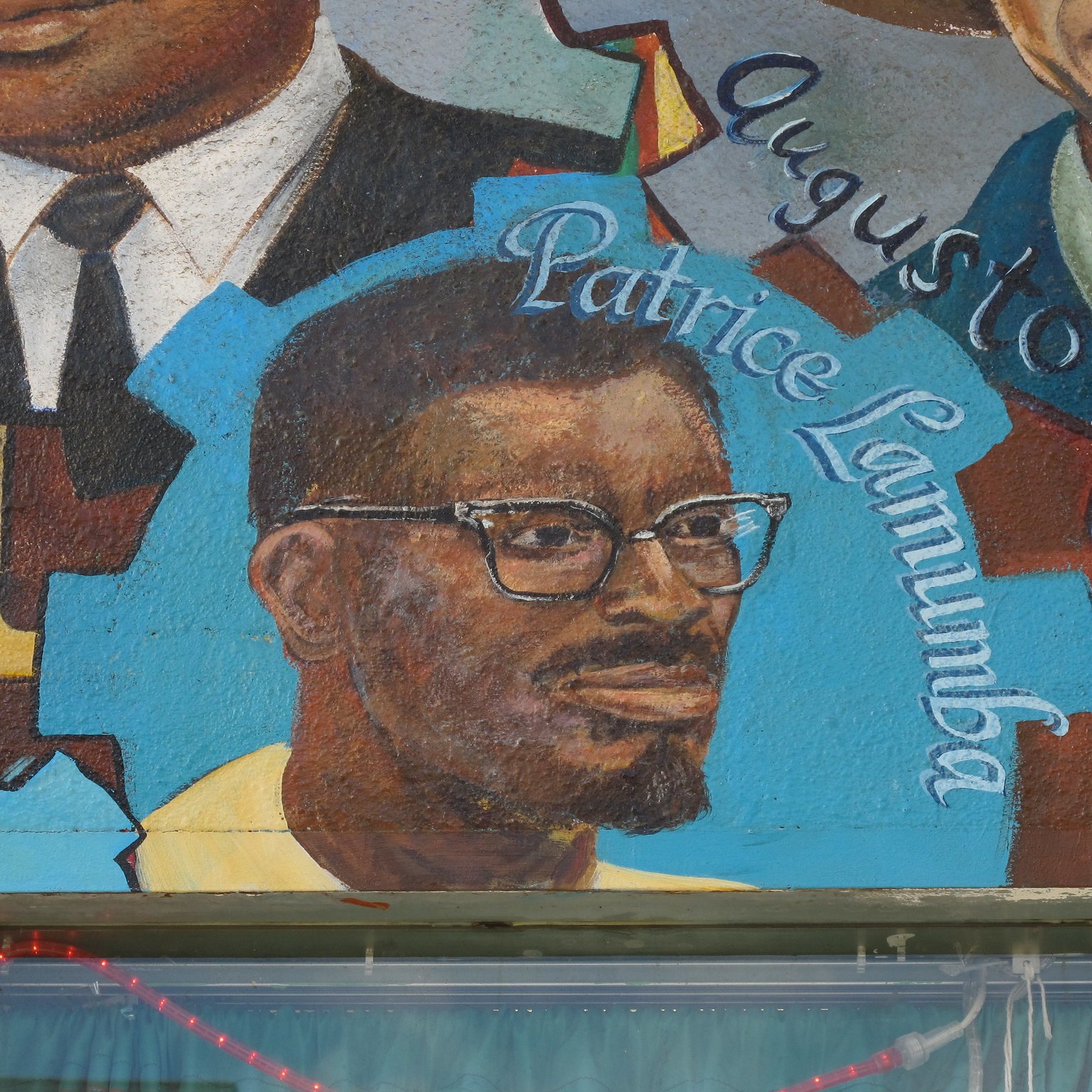 L'image semble représenter une fresque murale avec des portraits de différentes personnes. Au centre, on peut voir un homme portant des lunettes, avec une barbe et un sourire. Son nom, "Patrice Lumumba", est écrit en dessous de son portrait. Les autres éléments de la fresque peuvent inclure des symboles ou des motifs colorés, contribuant à une ambiance artistique et culturelle. La fresque pourrait célébrer des figures historiques ou politiques importantes.